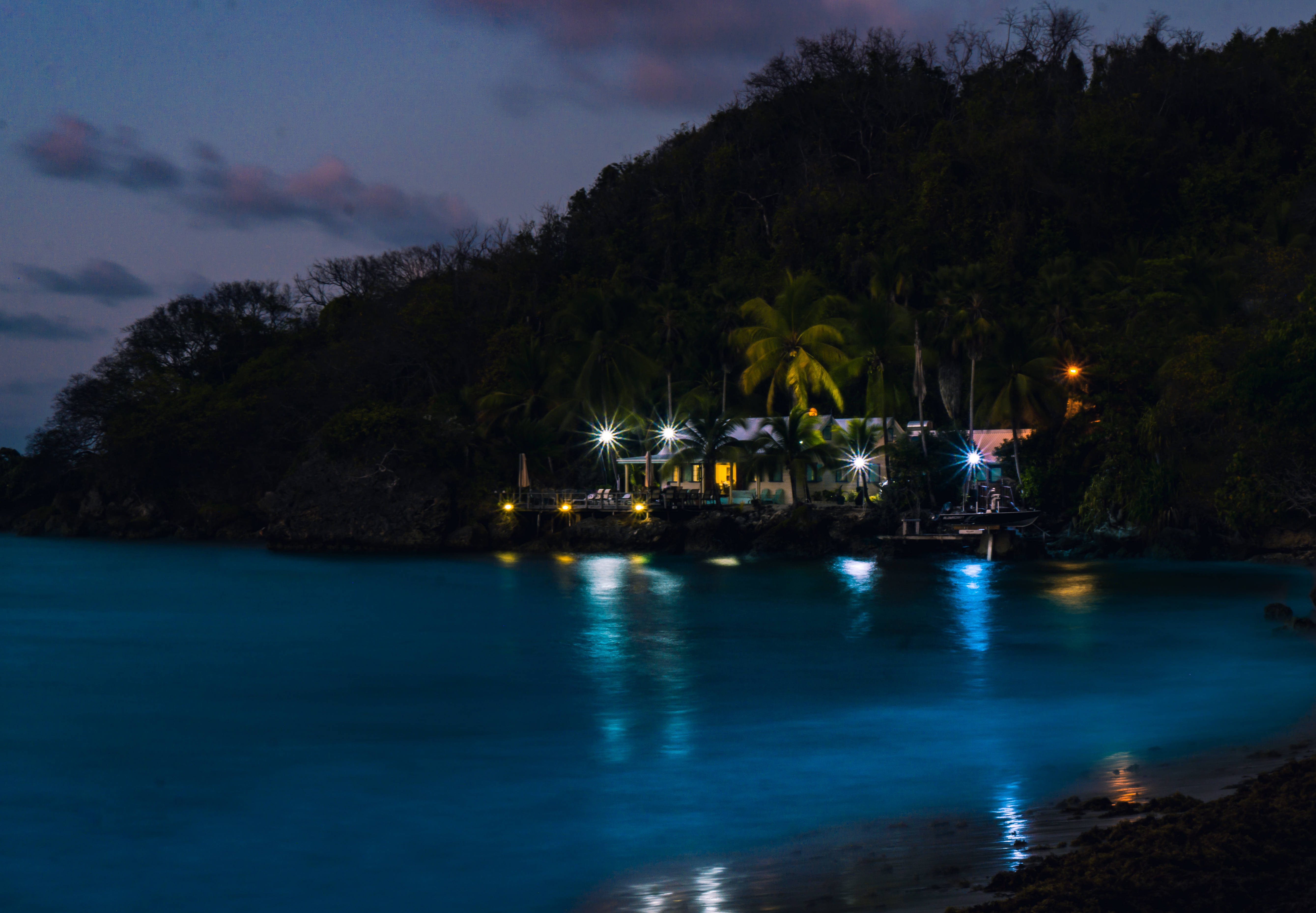 Barbados 桌布 壁紙 — 黃昏時分寧靜的熱帶海灘，一間亮燈的海邊餐廳在黑暗的山丘和寧靜的藍色海水映襯下閃耀。