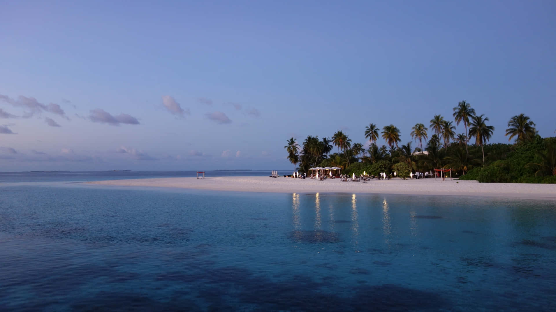 1920x1080 HD Beach 桌布 壁紙 — 黃昏時分寧靜的熱帶海灘，碧綠的海水、白色的沙灘、棕櫚樹和度假村的燈光在岸邊柔和地閃爍。