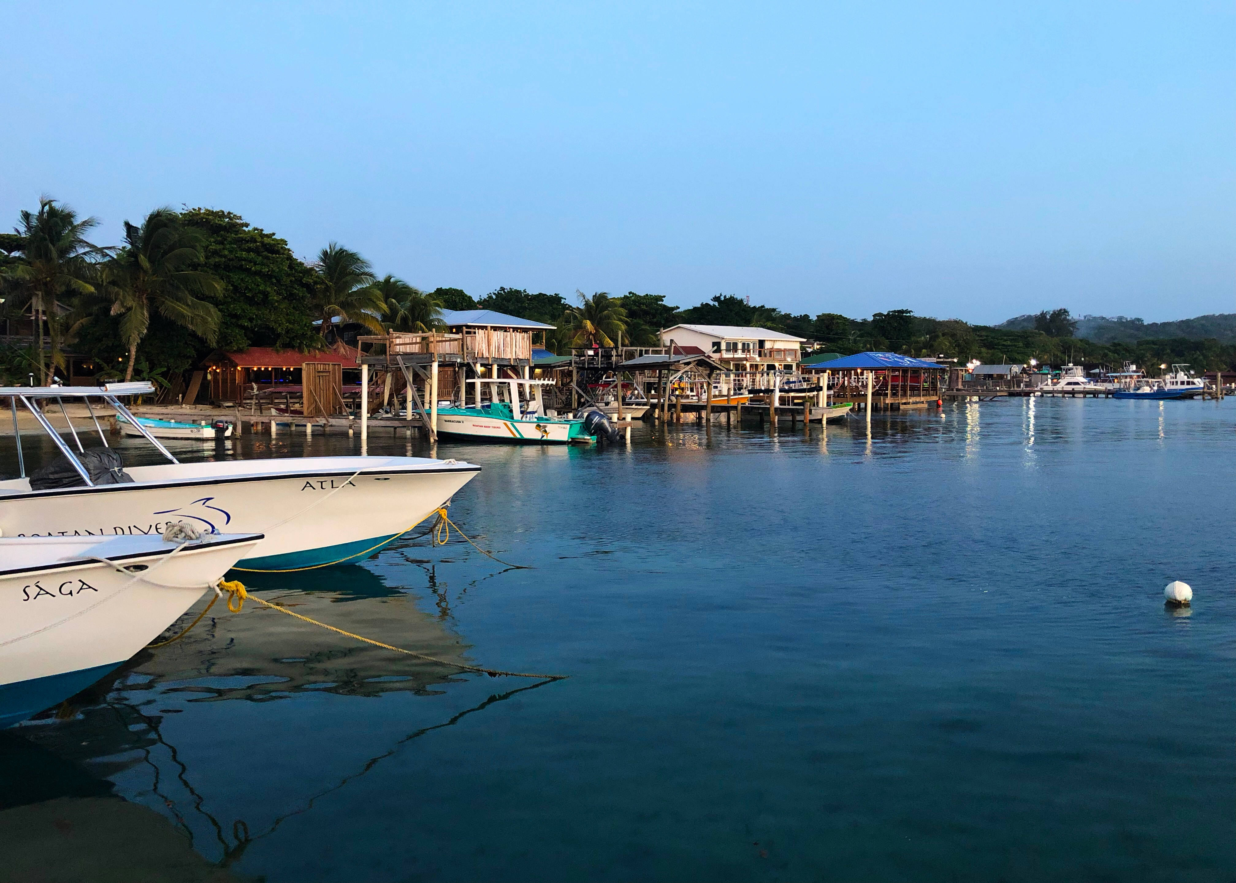 Honduras 桌布 壁紙 — 黃昏時分寧靜的熱帶港口，停泊的潛水船和高腳屋在平靜的碧綠海水中倒映。