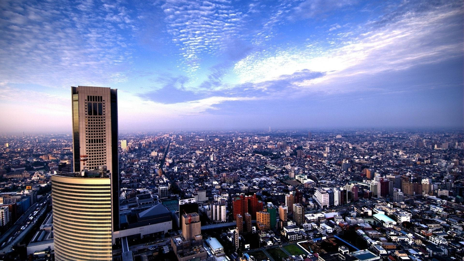 View 桌布 壁紙 — 黃昏時分廣闊城市景觀的鳥瞰圖，一座高聳的摩天大樓在前景中佔據主導地位，下方是戲劇性的藍天和縷縷雲彩。