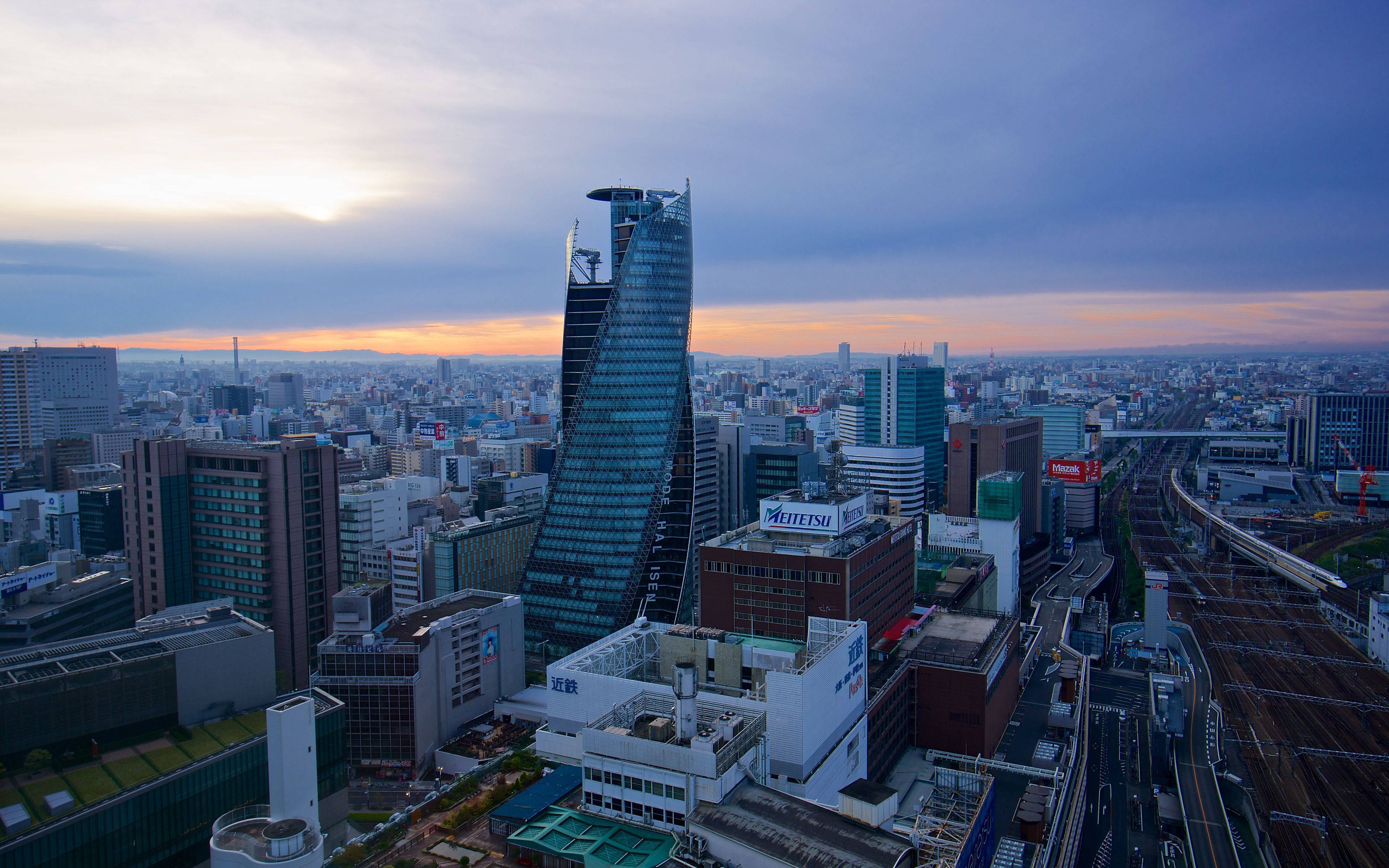 Nagoya 桌布 壁紙 — 黃昏時分廣闊城市景觀的鳥瞰圖，一座獨特的彎曲摩天大樓在漸變天空下主導天際線。
