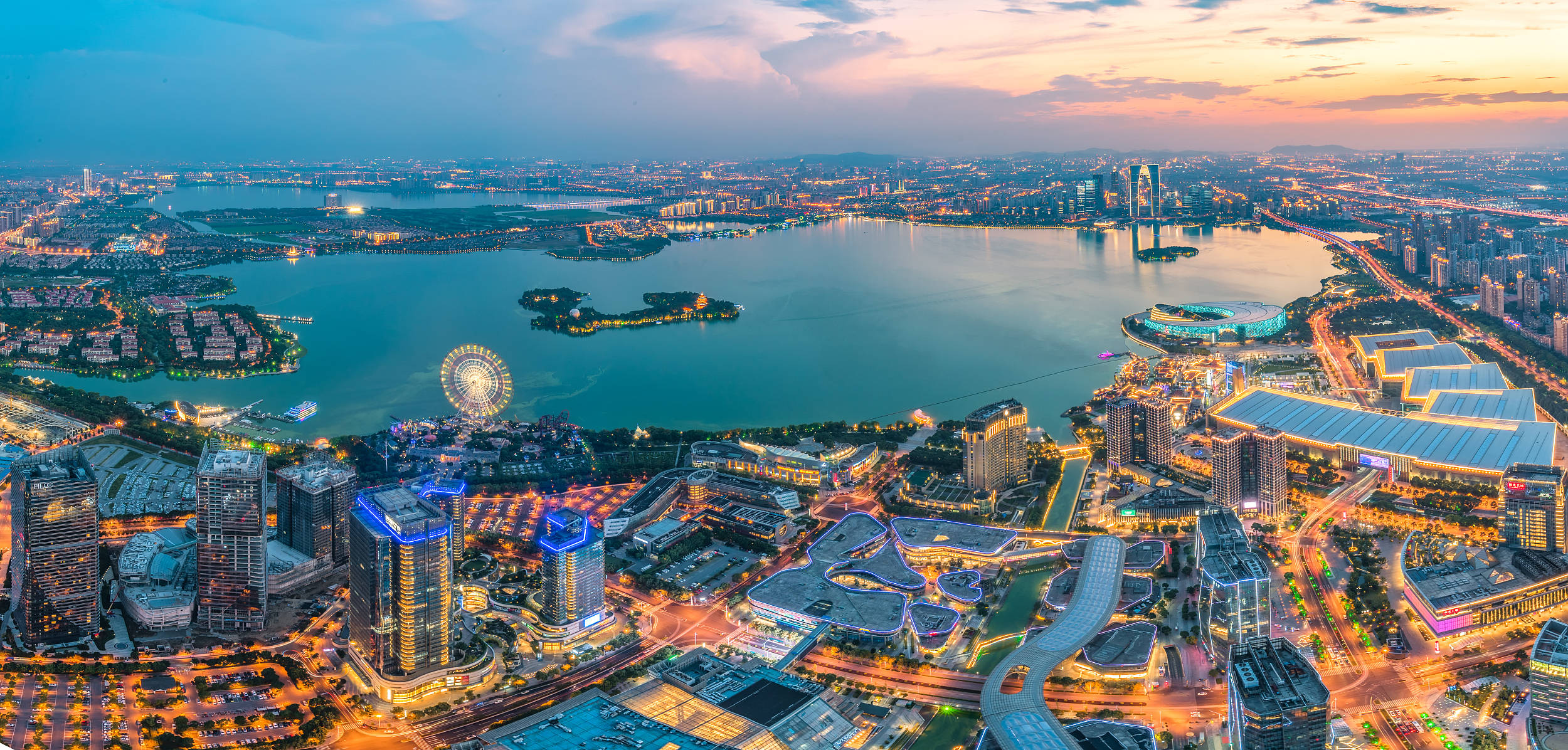 Suzhou 桌布 壁紙 — 黃昏時分廣闊城市景觀的鳥瞰圖，閃爍的摩天大樓和一個大型湖泊，設有摩天輪和燈光璀璨的島嶼。