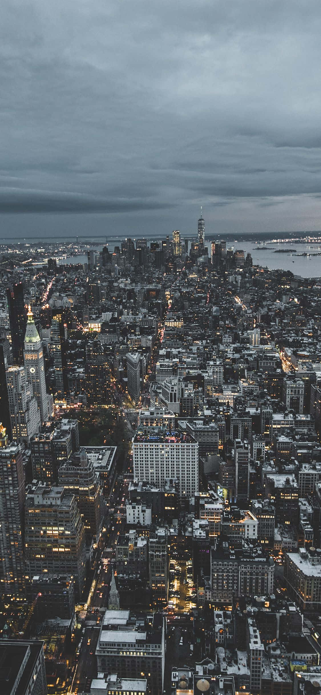iPhone Xs New York 桌布 壁紙 — 黃昏時分廣闊城市景觀的鳥瞰圖，陰沉灰暗的天空下閃爍的街燈和摩天大樓。