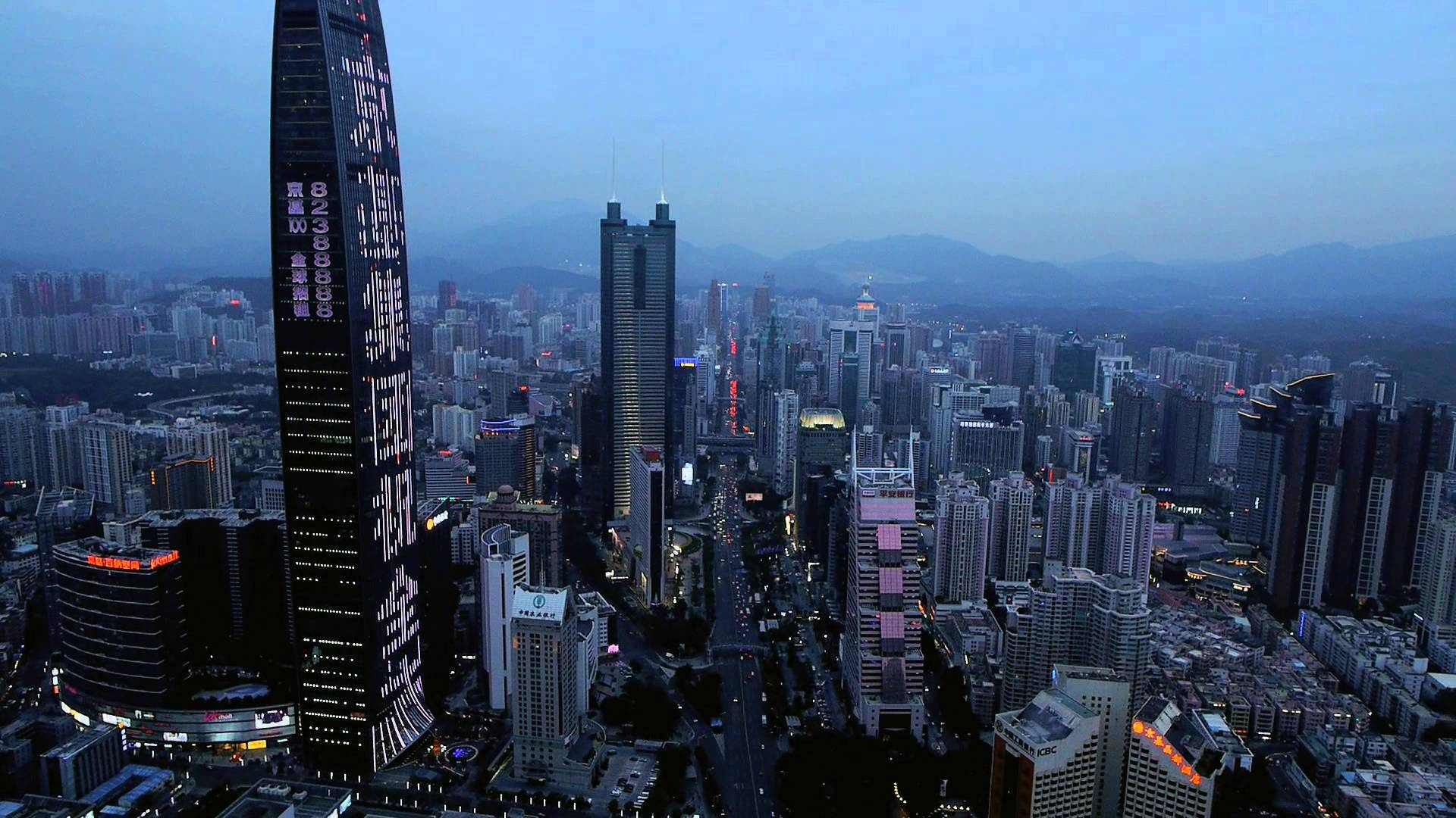 Shenzhen 桌布 壁紙 — 黃昏時分廣闊城市景觀的鳥瞰圖，高聳的摩天大樓在漸暗的藍天和遠山映襯下亮起燈光。