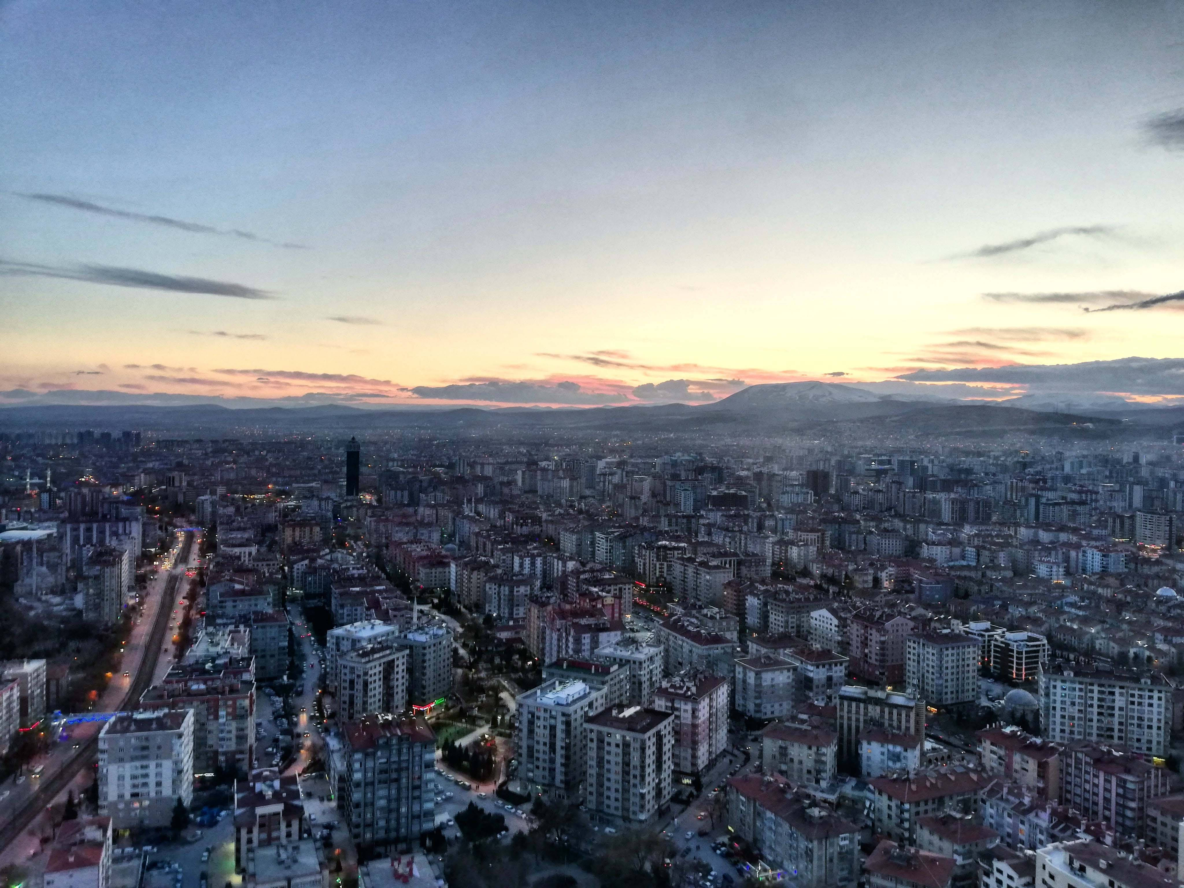Konya 桌布 壁紙 — 黃昏時分廣闊的城市景觀，溫暖的日落色調融入密集住宅樓和遠處山脈上方清涼的藍色天空。