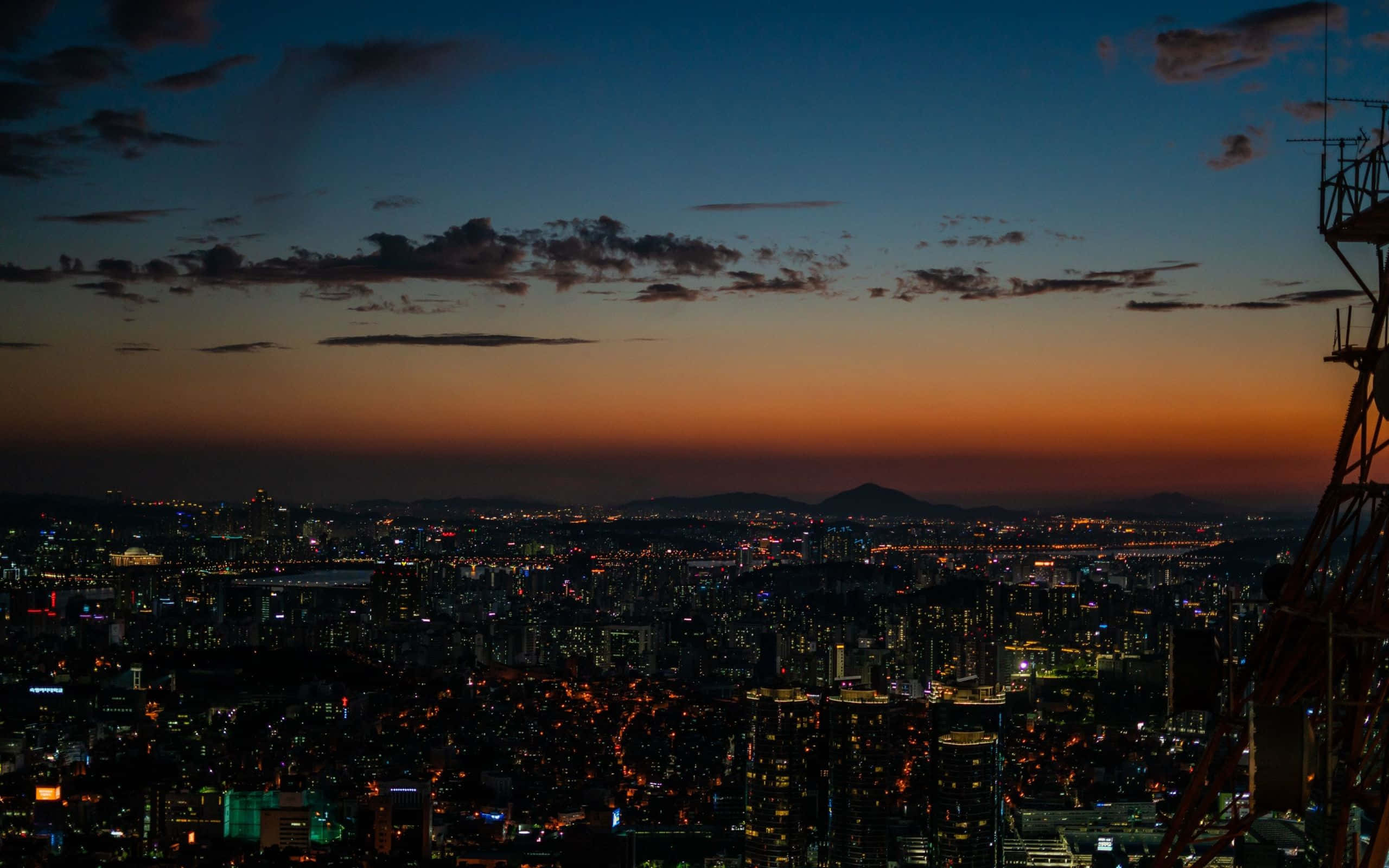 korean 桌布 壁紙 — 黃昏時分廣闊的城市景觀，溫暖的橙色地平線逐漸消失在深藍色的天空中，城市燈光閃爍。