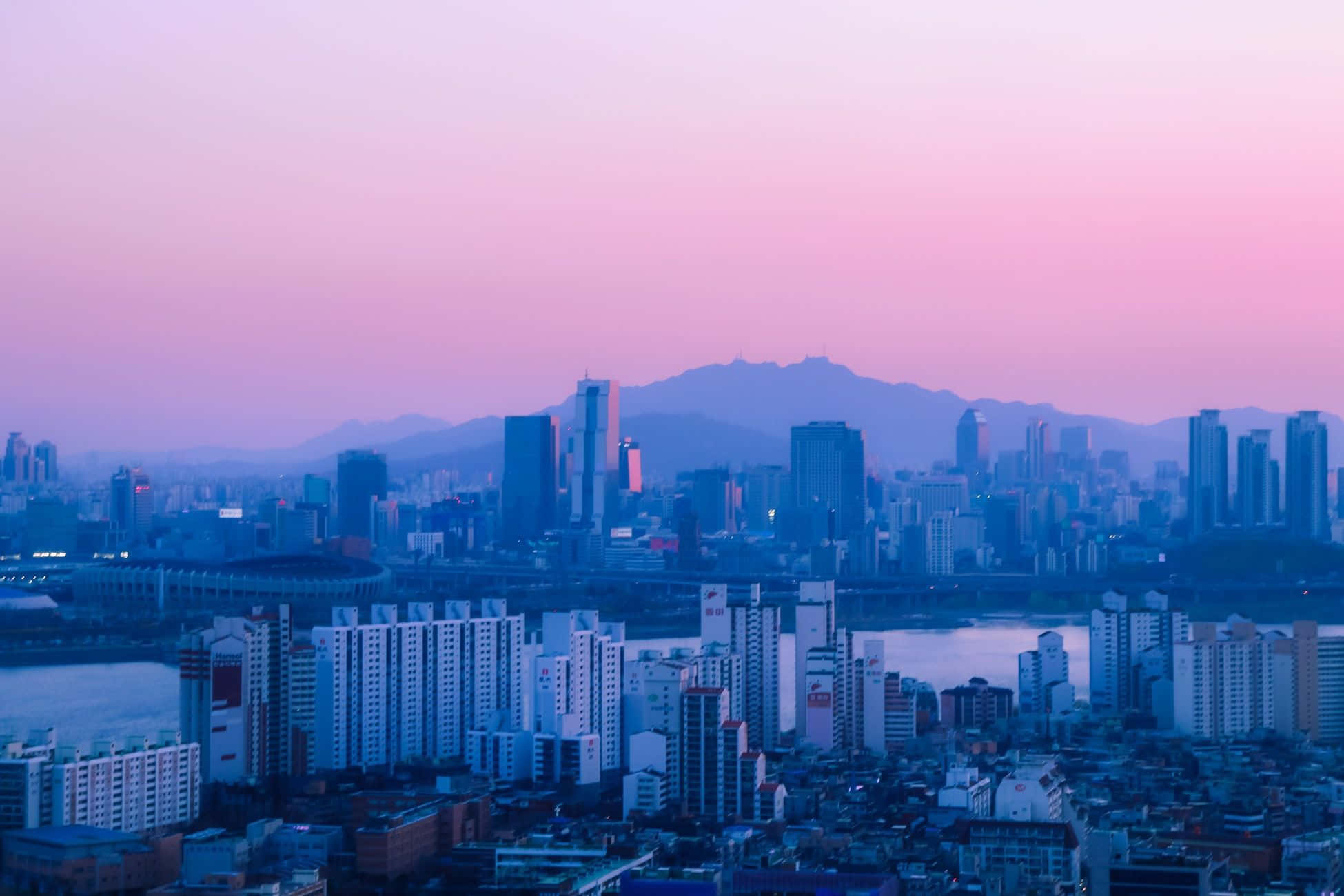 Aesthetic Korean 桌布 壁紙 — 黃昏時分廣闊的城市景觀，現代摩天大樓和遠處山脈上方呈現粉紅色和紫色的天空。