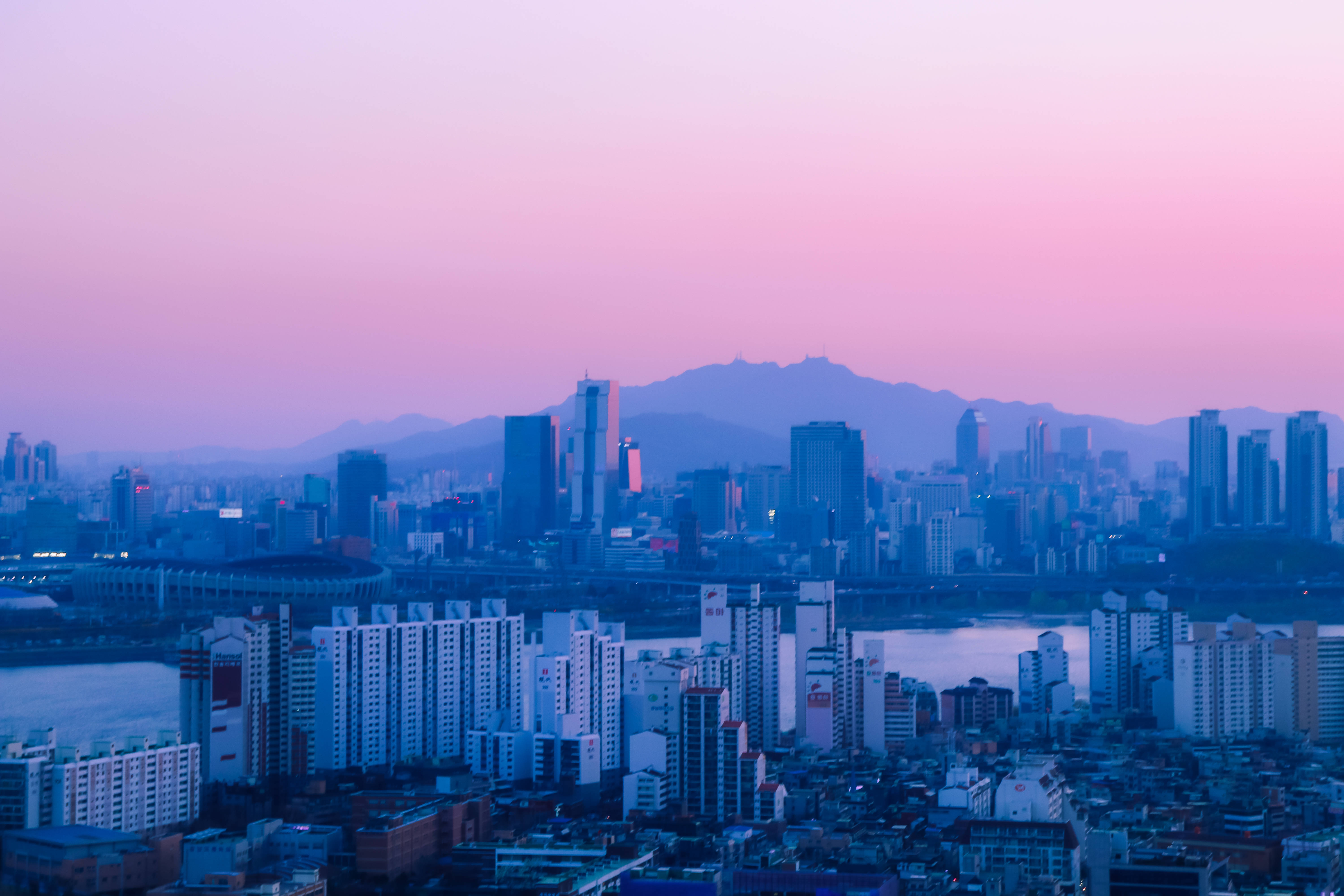 動漫 桌布 壁紙 — 黃昏時分廣闊的城市景觀，粉紅色和紫色的天空籠罩著現代摩天大樓和遠處的山脈。