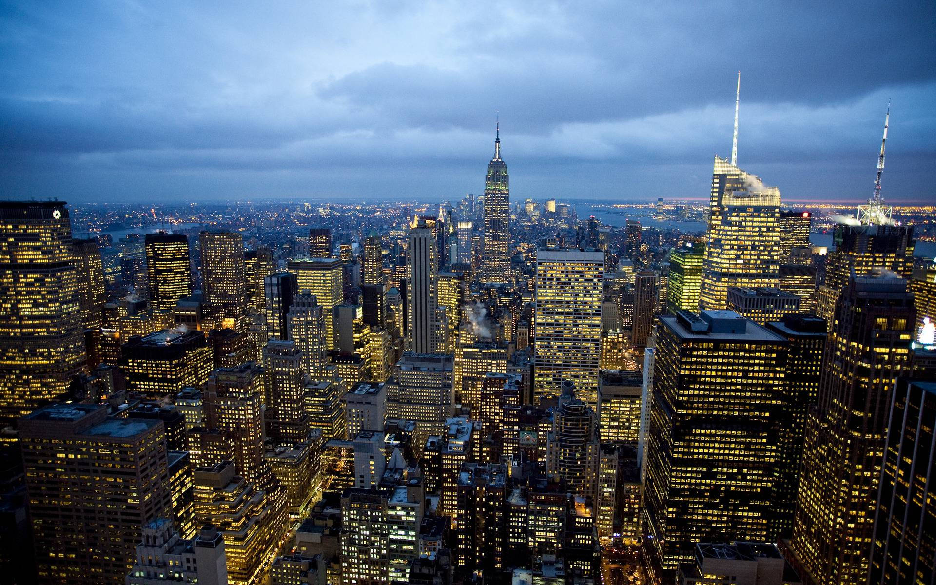 New York City Desktop 桌布 壁紙 — 黃昏時分廣闊的城市景觀，高聳的摩天大樓在憂鬱的藍天下溫暖地閃耀。