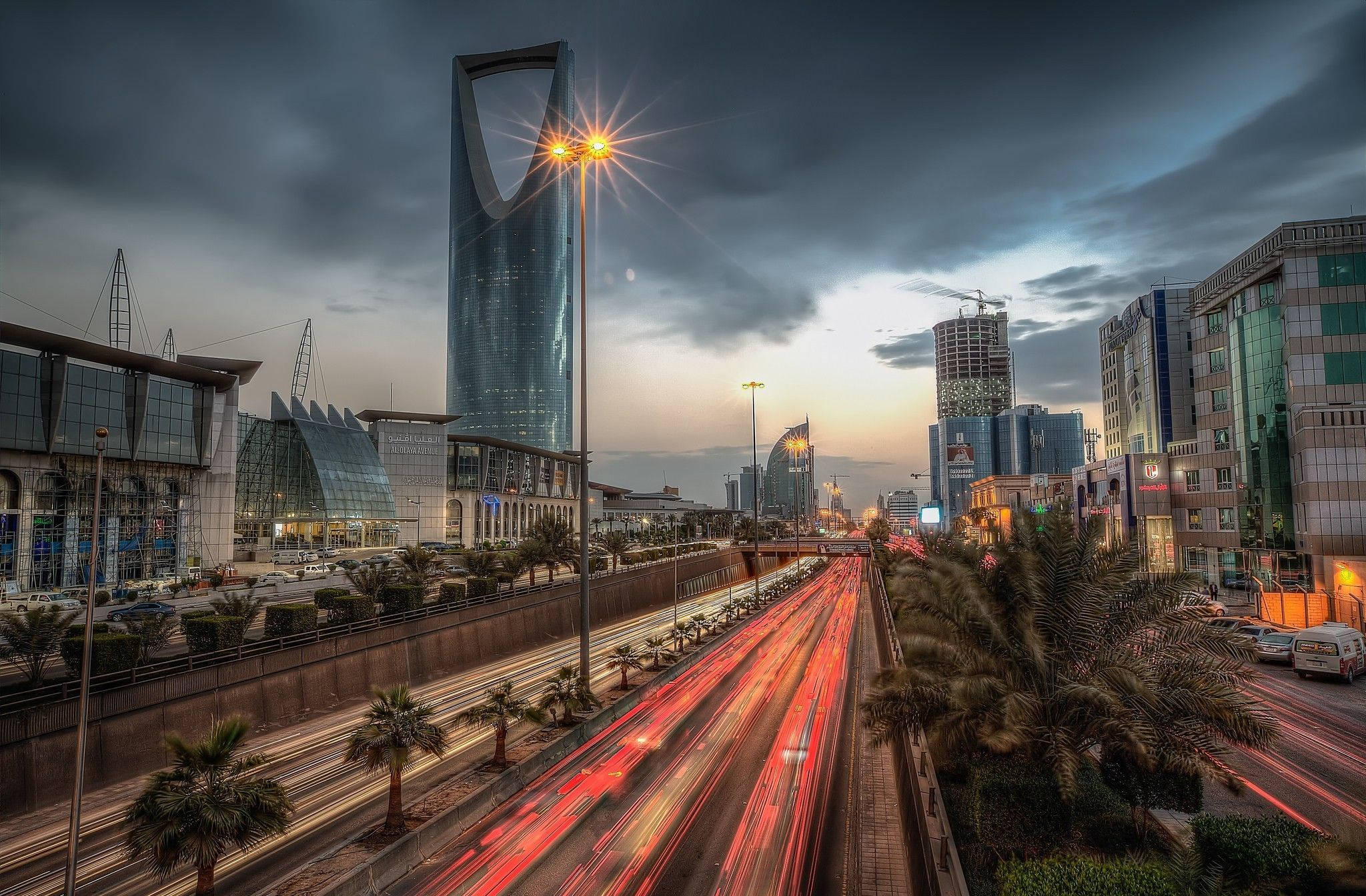 Saudi 桌布 壁紙 — 黃昏時分戲劇性的城市天際線，帶有紅色車流燈光軌跡，標誌性的王國塔刺穿多雲的天空。