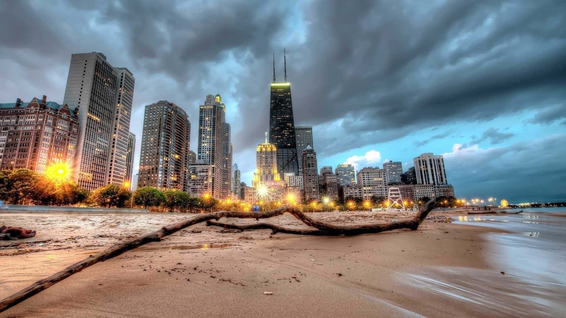 1920x1080 HD Beach 桌布 壁紙 — 黃昏時分戲劇性的城市海灘場景，前景有倒下的圓木，芝加哥天際線在暴風雲下被金色路燈照亮。