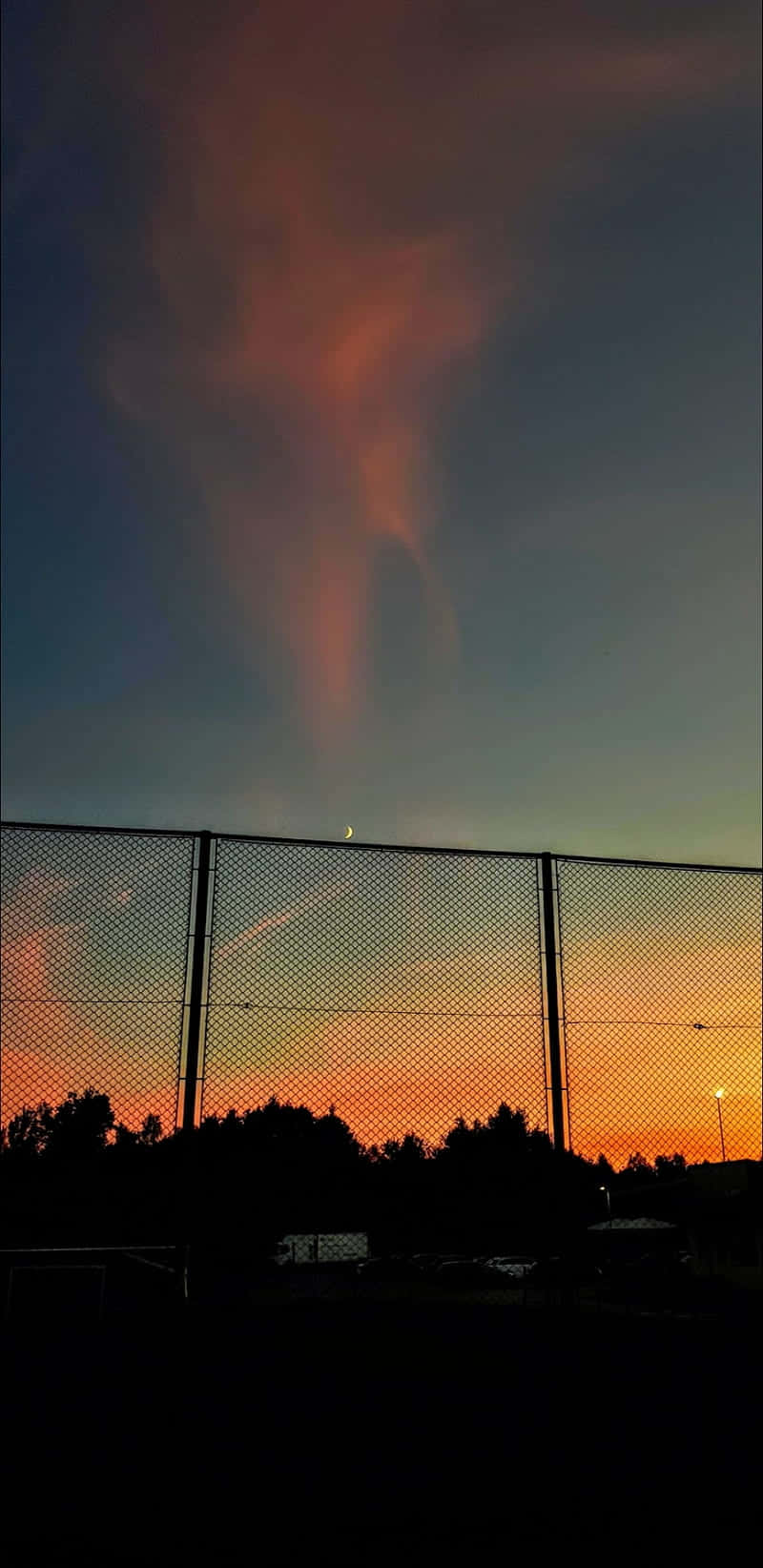 桌布 壁紙 — 黃昏時分戲劇性的天空，火紅的橙色雲朵和一彎新月懸掛在深色鐵絲網圍欄上方。