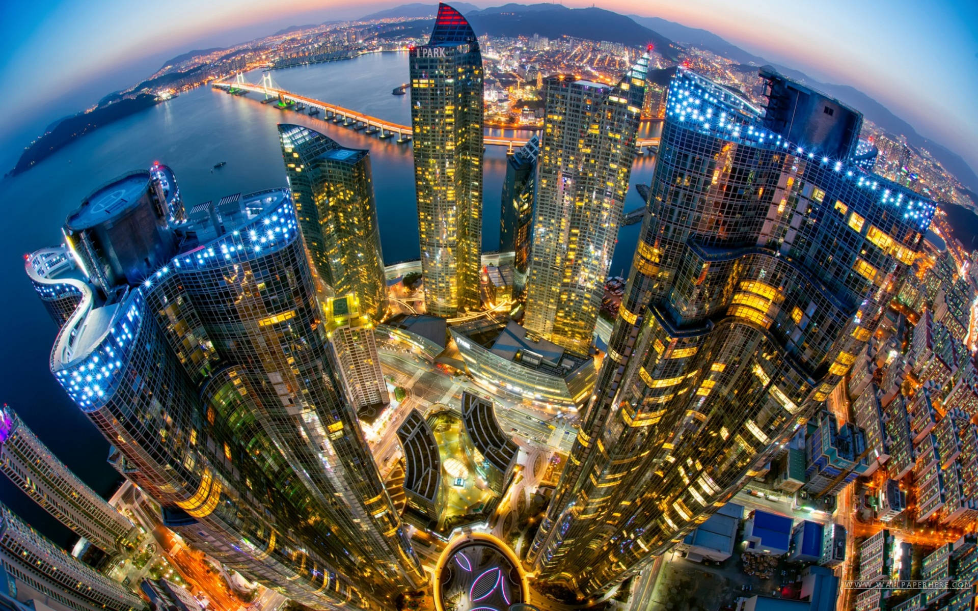 Busan 桌布 壁紙 — 黃昏時分未來城市景觀的魚眼視角，發光的摩天大樓和水上的橋樑。