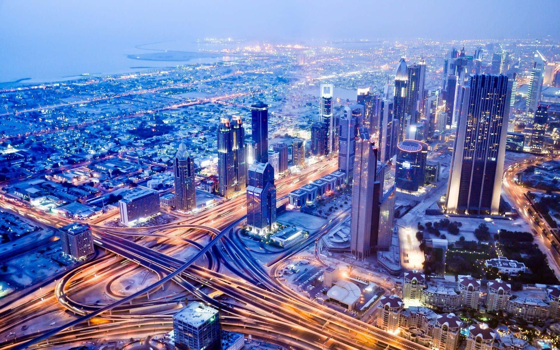 Uae 桌布 壁紙 — 黃昏時分未來城市景觀的鳥瞰圖，閃爍的摩天大樓和照明高速公路蜿蜒穿過密集的城市發展。