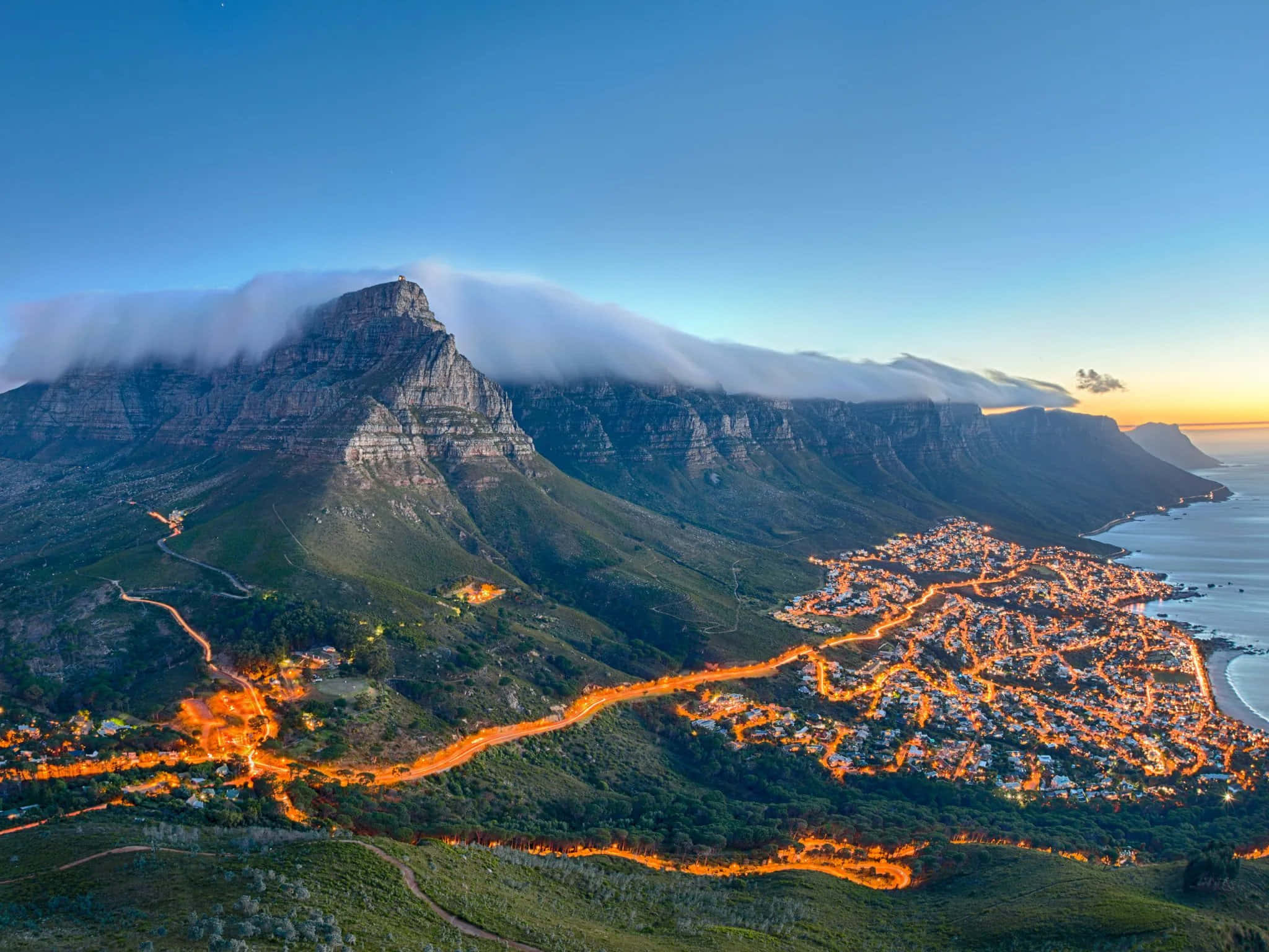 South Africa 桌布 壁紙 — 黃昏時分桌山的鳥瞰圖，發光的城市燈光蜿蜒穿過綠色山丘和海洋海岸線。
