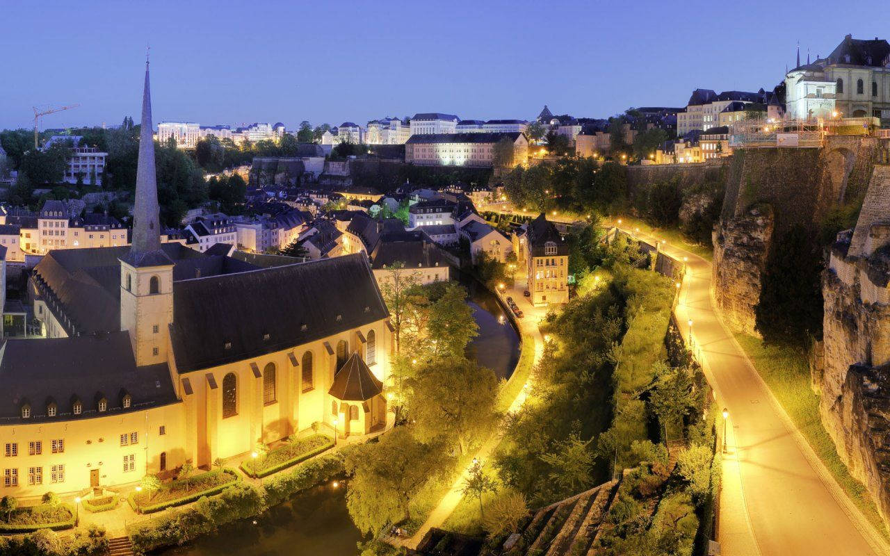 Luxembourg 桌布 壁紙 — 黃昏時分歷史悠久的歐洲小鎮鳥瞰圖，溫暖燈光照亮的教堂和蜿蜒的街道依偎在懸崖邊。