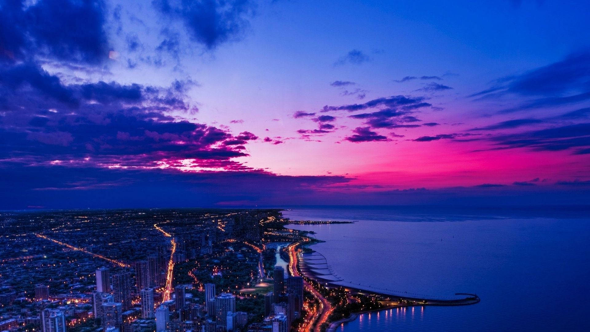 View 桌布 壁紙 — 黃昏時分沿海城市的鳥瞰圖，充滿活力的粉紅色和紫色天空在發光的街道和寧靜的水面上方。