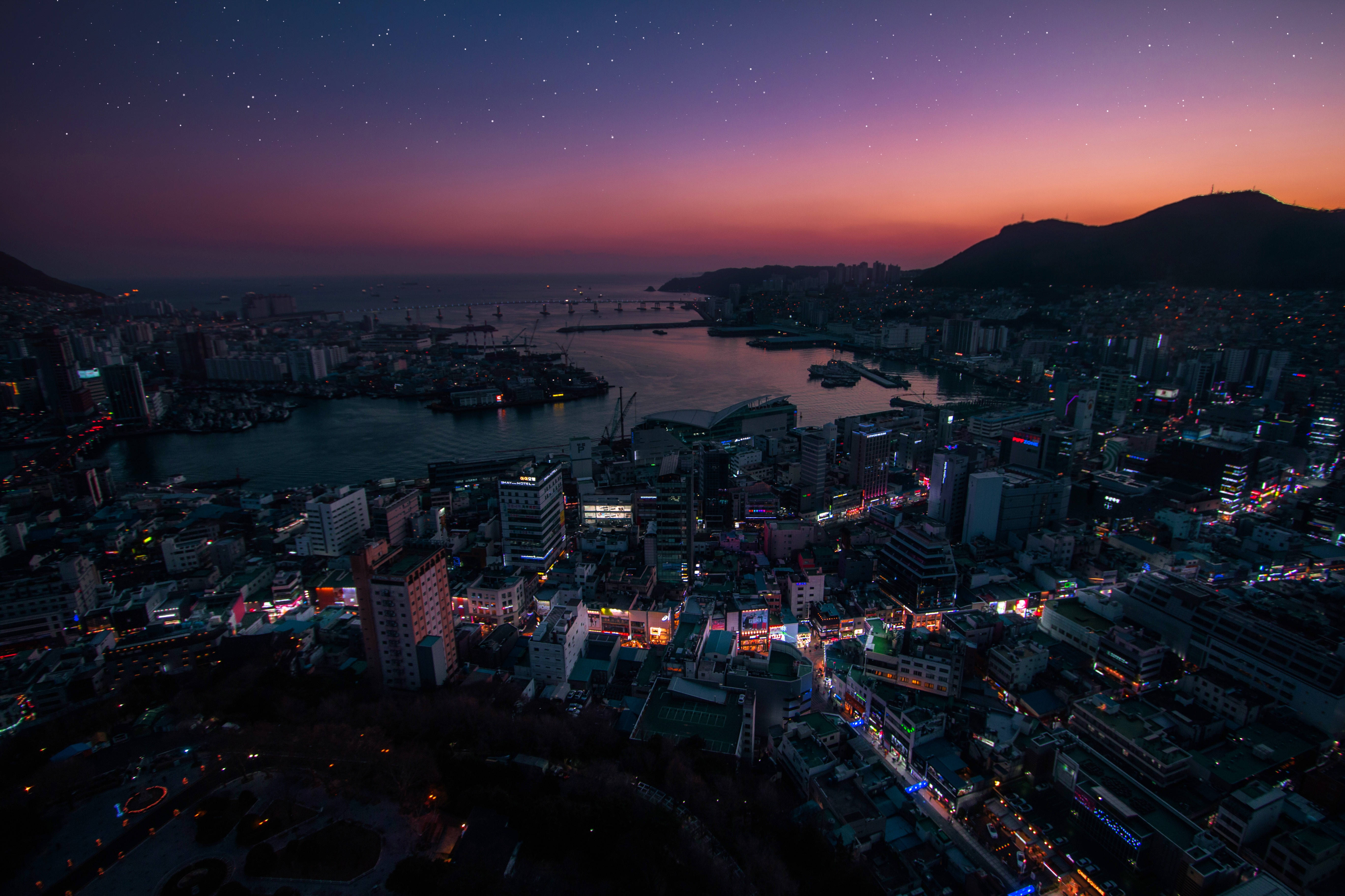 南韓 桌布 壁紙 — 黃昏時分沿海城市的鳥瞰圖，發光的街燈和星星在紫橙色天空中浮現。
