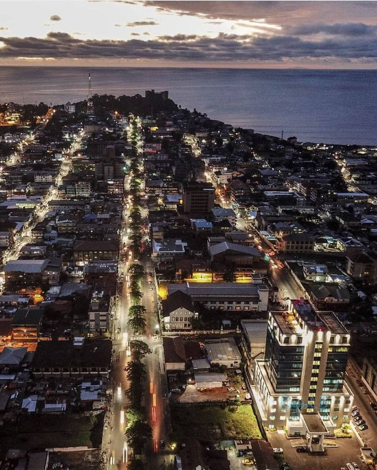 Liberia 桌布 壁紙 — 黃昏時分沿海城市的鳥瞰圖，街道燈光閃爍，建築物緊貼海岸線，天空多雲。