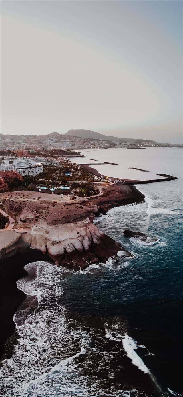 西班牙 桌布 壁紙 — 黃昏時分沿海小鎮的鳥瞰圖，海浪拍打著深色火山懸崖，遠處的山脈在柔和的粉彩天空下。