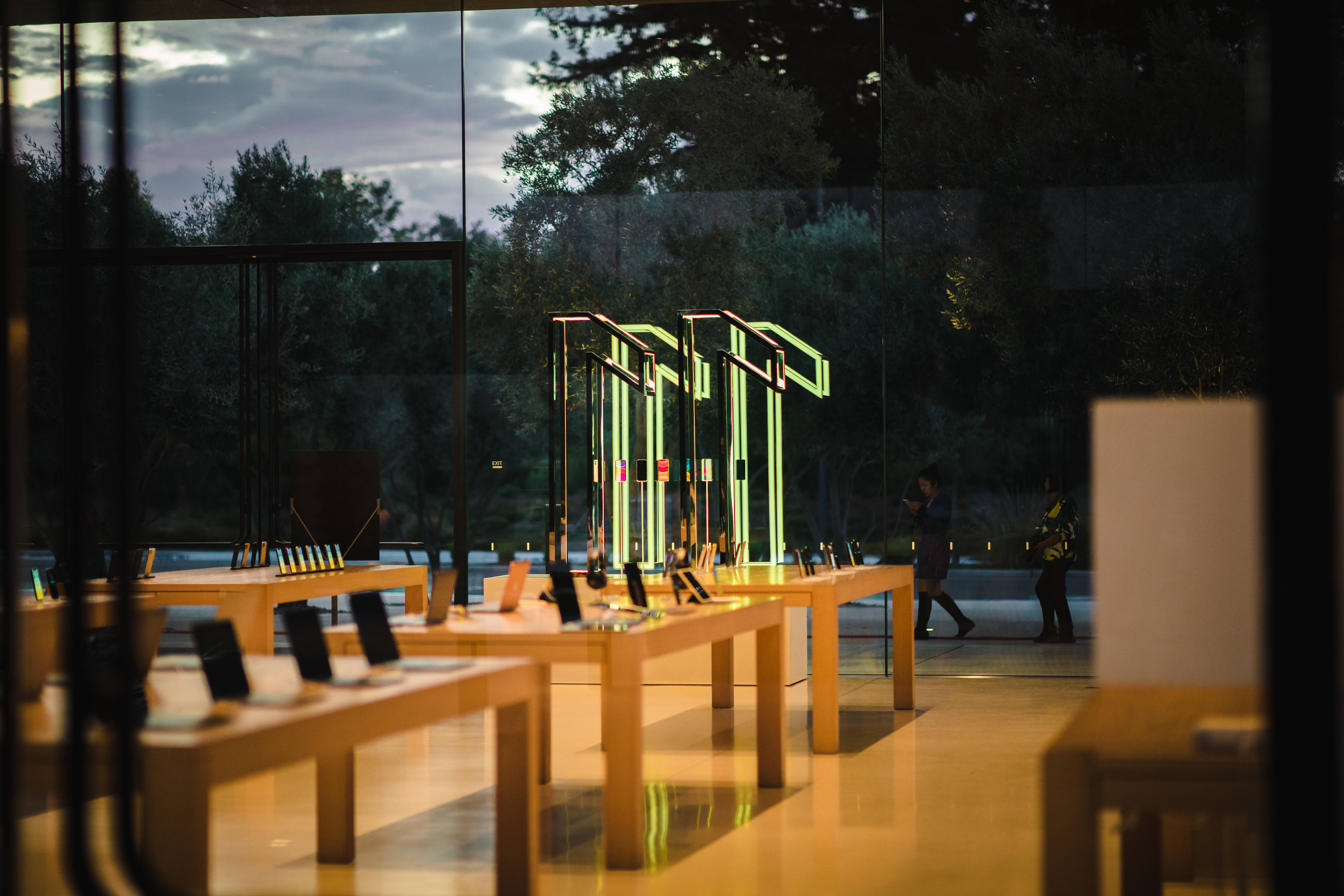 iPhone 11 桌布 壁紙 — 黃昏時分溫暖照明的Apple Store內部，發光的產品展示和玻璃牆外的剪影訪客。