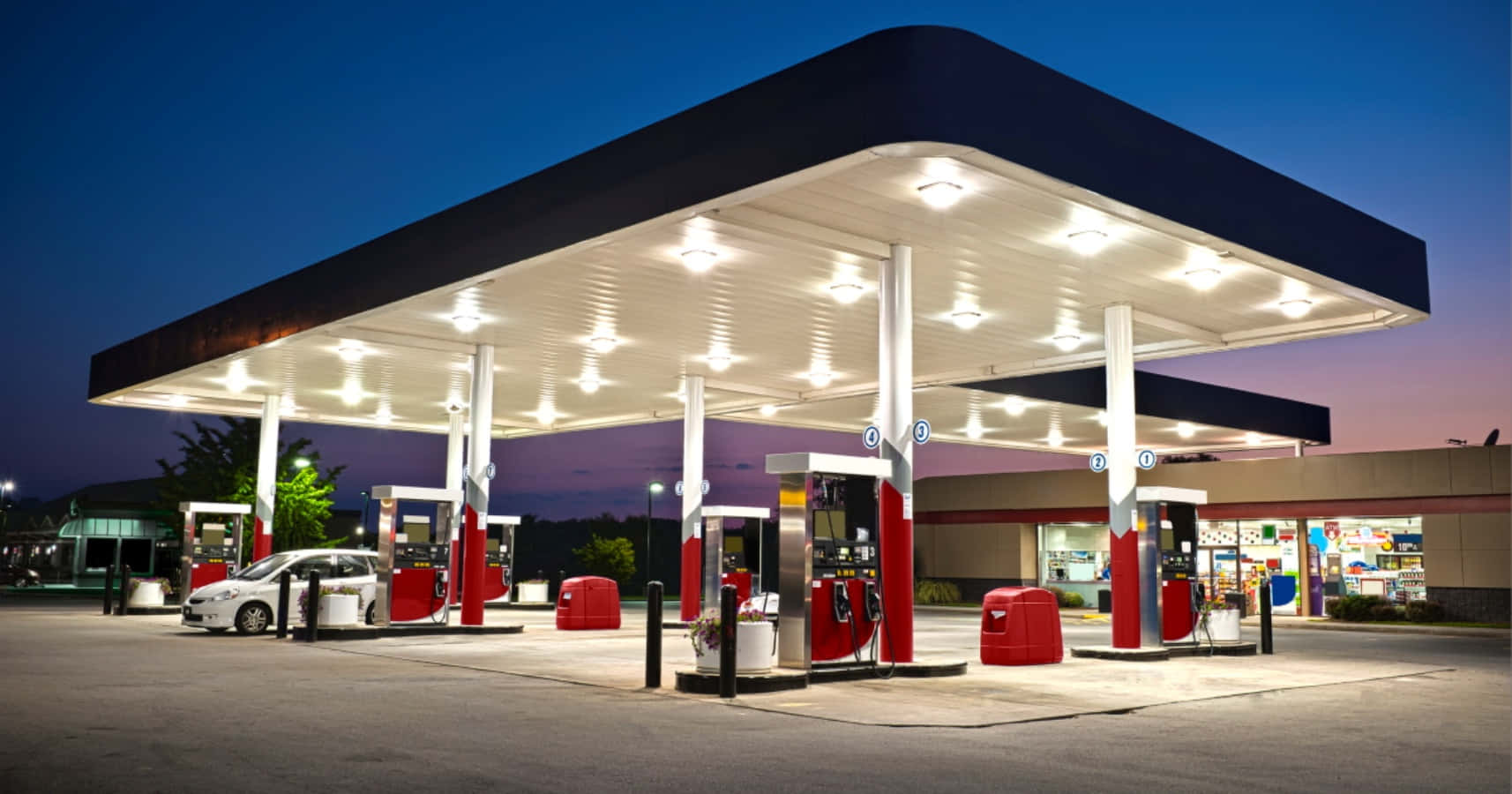 Gas Station 桌布 壁紙 — 黃昏時分燈火通明的加油站，一輛銀色汽車停泊在現代化遮雨棚下的油泵旁。