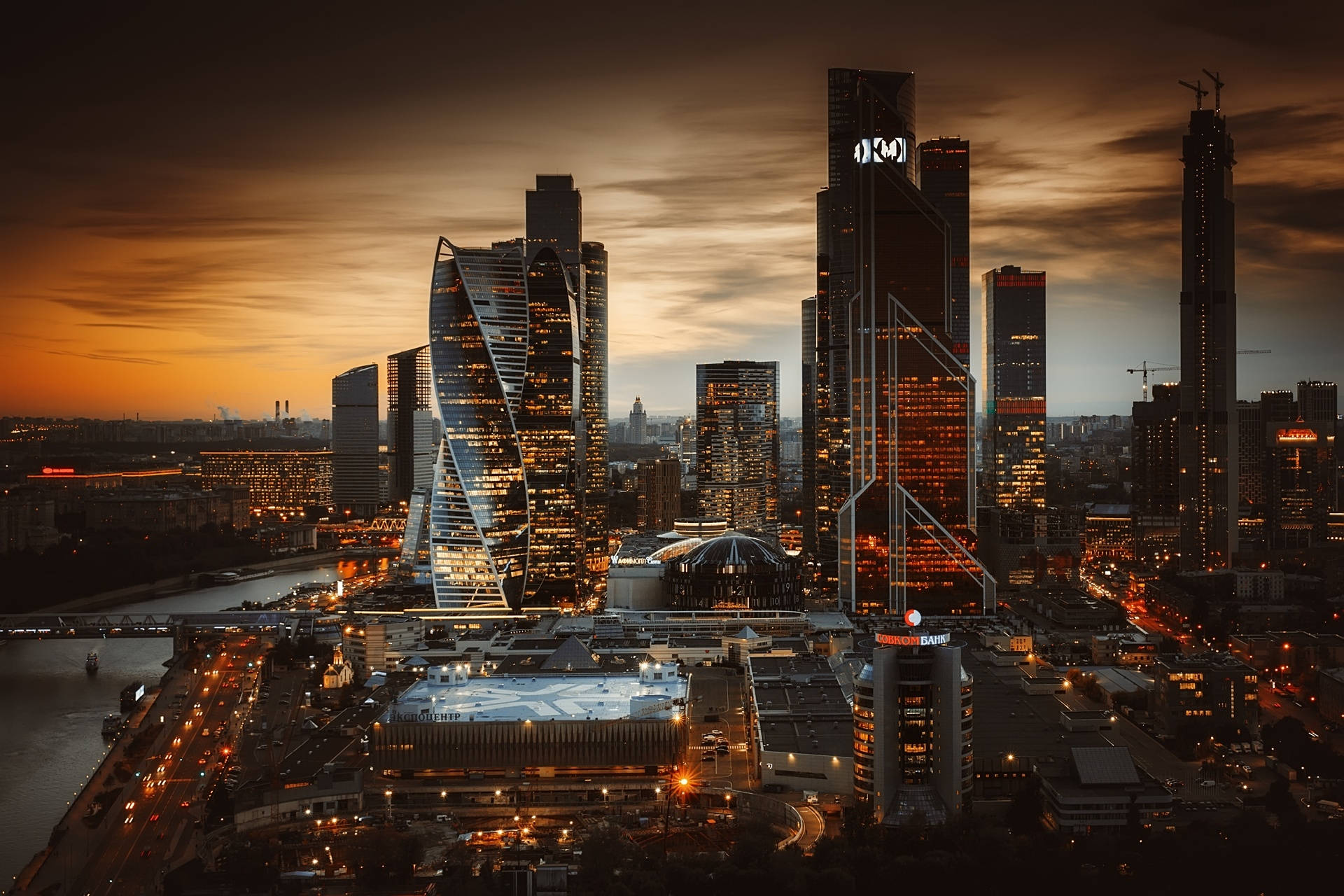 Russia 桌布 壁紙 — 黃昏時分現代城市天際線的鳥瞰圖，有燈火通明的摩天大樓和河流反射溫暖的橙色和金色色調。