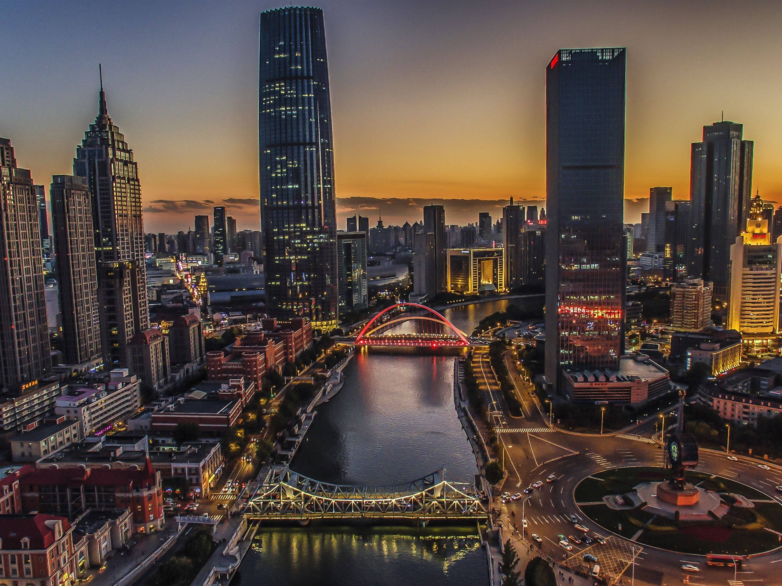 Tianjin 桌布 壁紙 — 黃昏時分現代城市天際線的鳥瞰圖，閃爍的摩天大樓和河上的一座紅色拱橋。