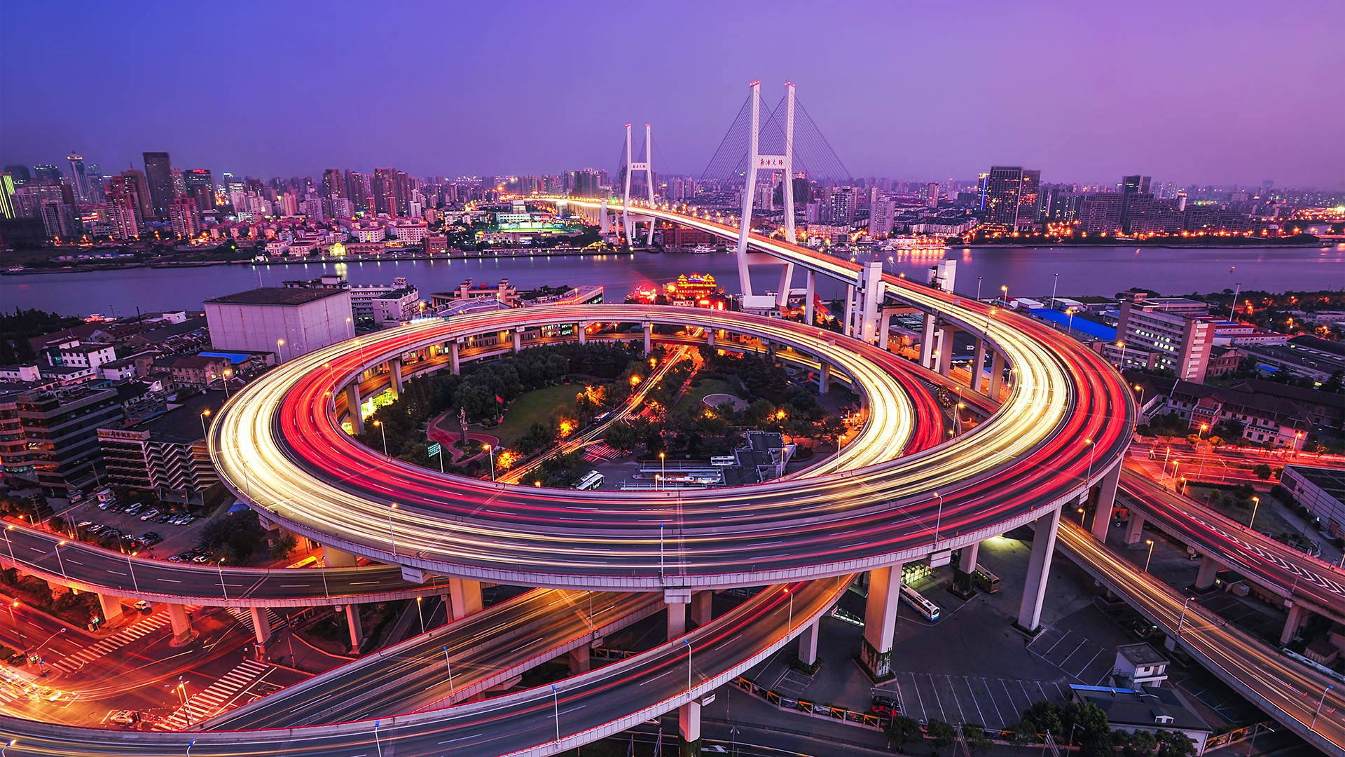 Shanghai 桌布 壁紙 — 黃昏時分發光的螺旋形高速公路交匯處的鳥瞰圖，帶有汽車燈光軌跡和背景中的城市天際線。