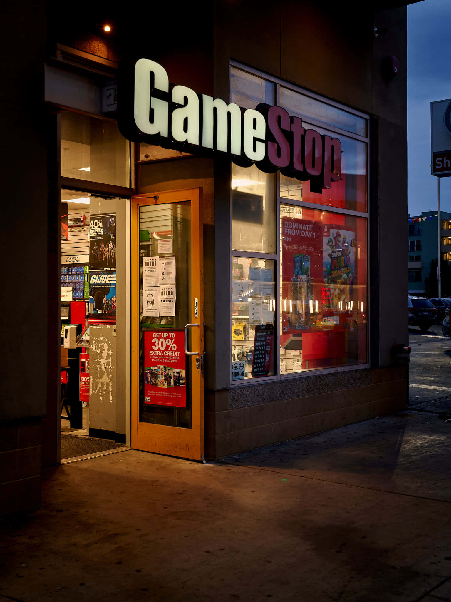GameStop 桌布 壁紙 — 黃昏時分的GameStop商店入口，溫暖的室內燈光透過玻璃窗和木門閃爍。