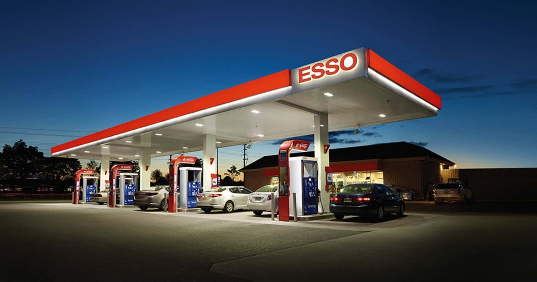 Petrol Station 桌布 壁紙 — 黃昏時分的埃索加油站，汽車在明亮的紅白簷篷下加油，背景是深藍色的天空。
