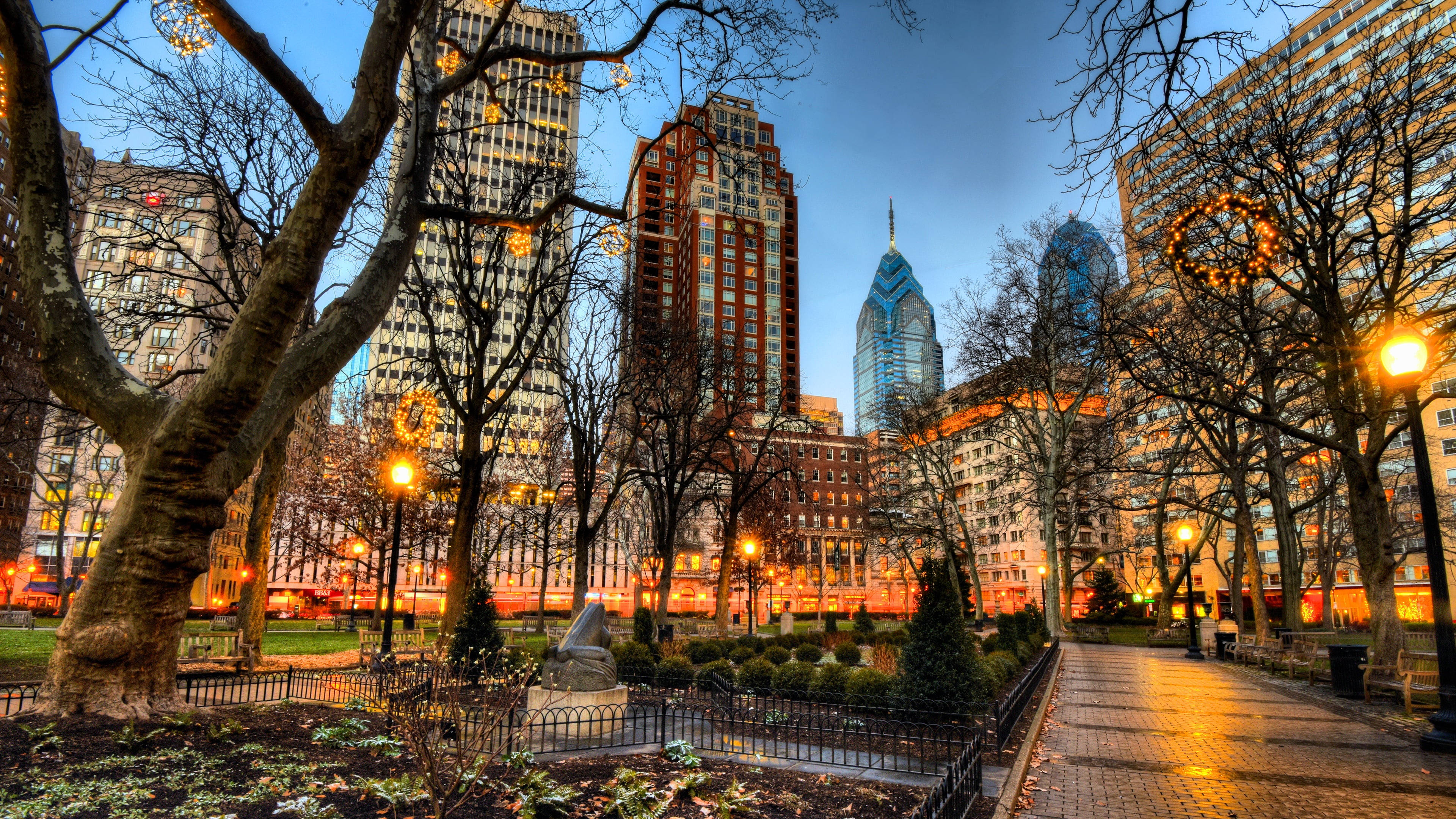 Philadelphia 桌布 壁紙 — 黃昏時分的城市公園，光禿禿的樹木在溫暖的路燈下閃耀，背景是高聳的摩天大樓。