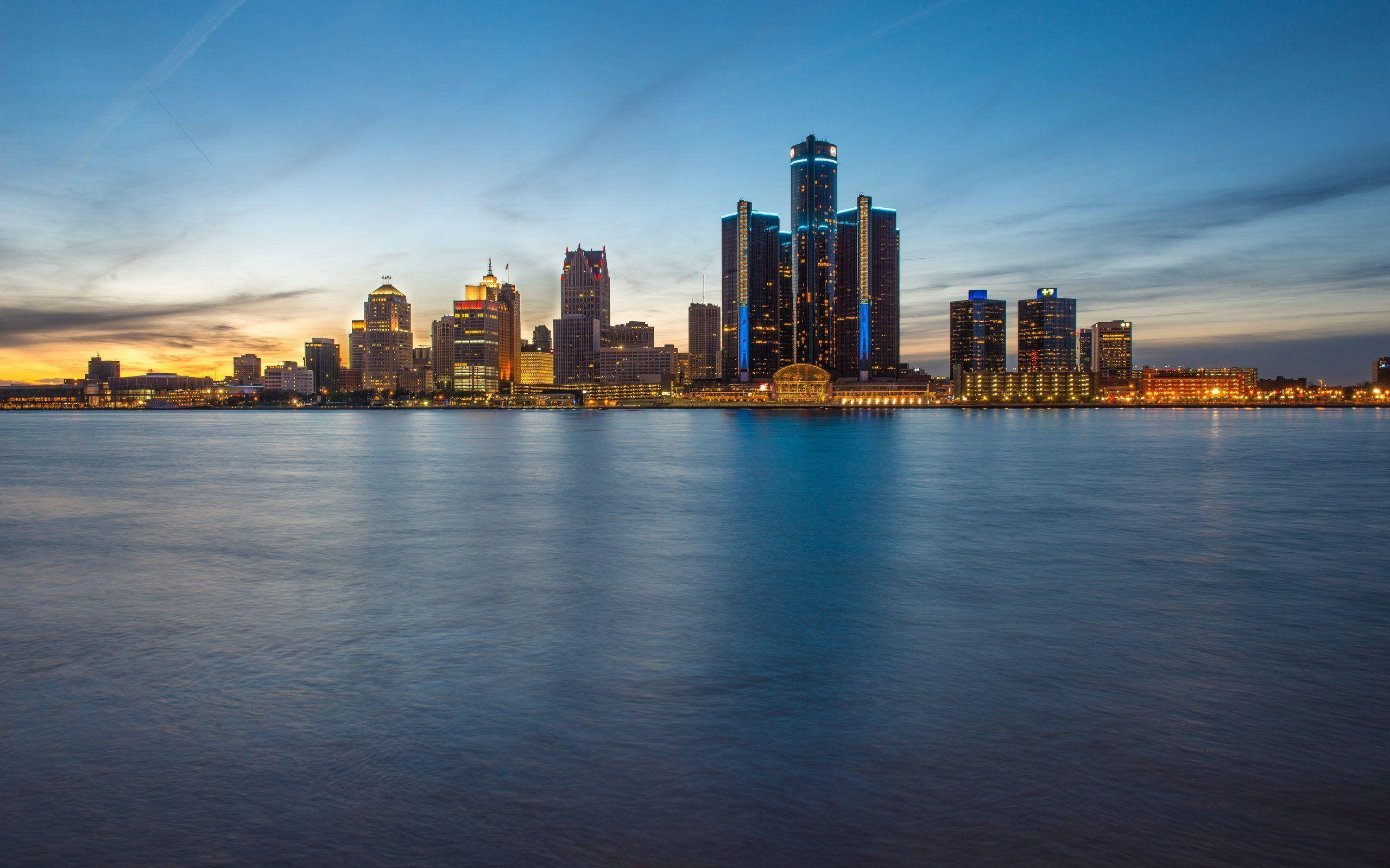 detroit 桌布 壁紙 — 黃昏時分的城市天際線，在漸變天空下，寧靜水面倒映著燈火通明的摩天大樓。