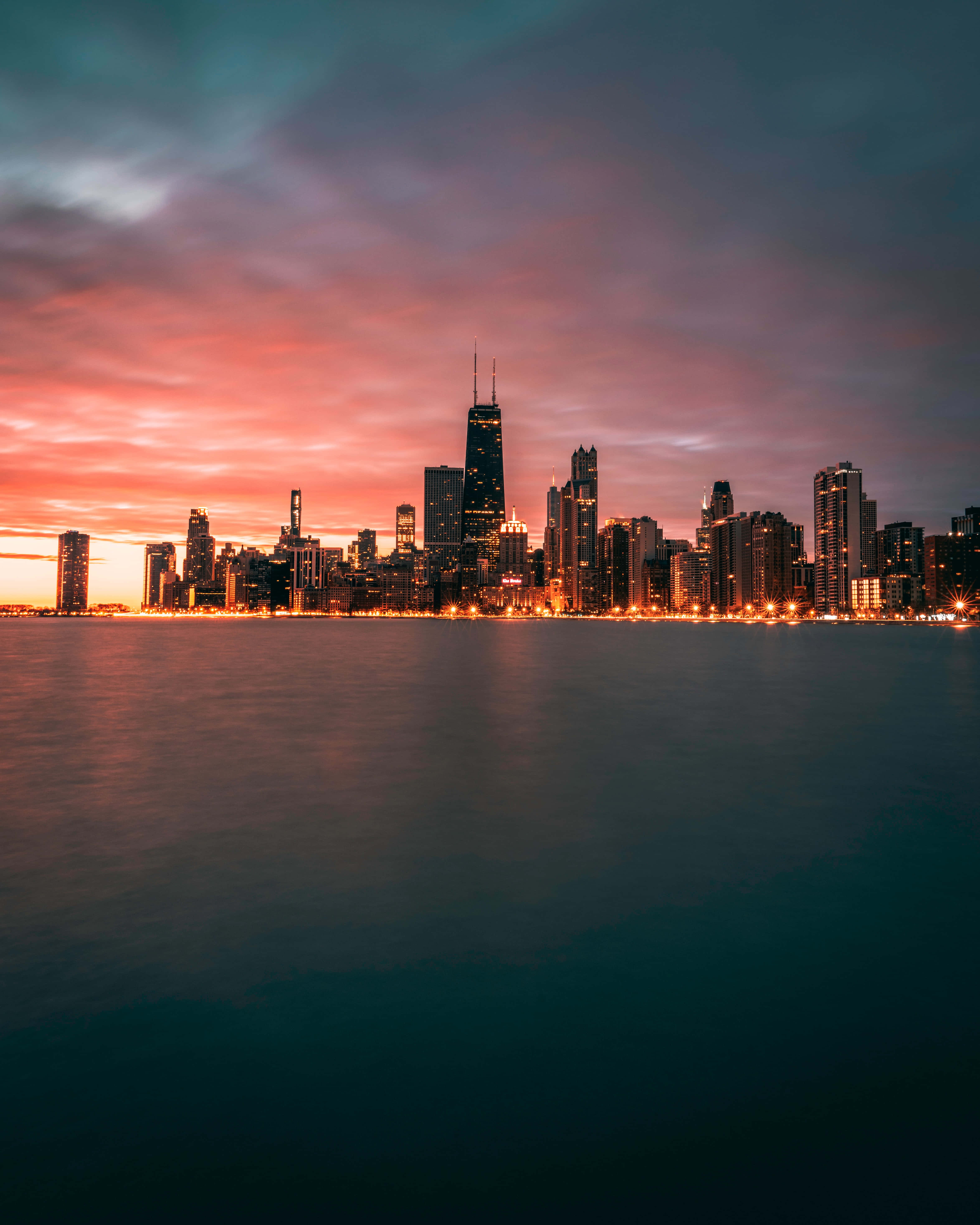 Chicago City Night 桌布 壁紙 — 黃昏時分的城市天際線，溫暖的橙色燈光映照在平靜的水面上，天空呈現漸變色。