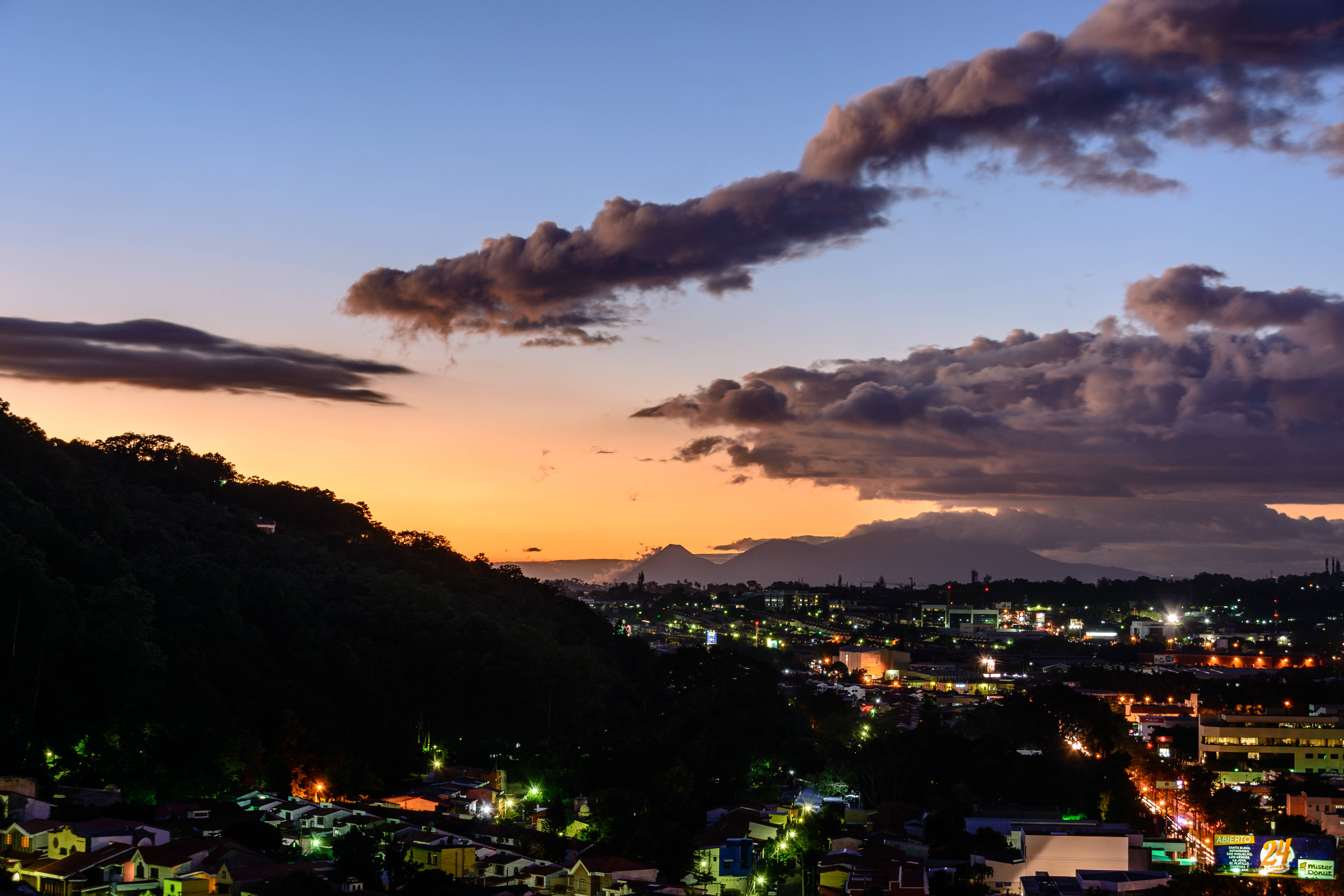 El Salvador 桌布 壁紙 — 黃昏時分的城市天際線，溫暖的橙色夕陽餘暉在烏雲後閃耀，照亮了廣闊的城市區域，燈光閃爍。