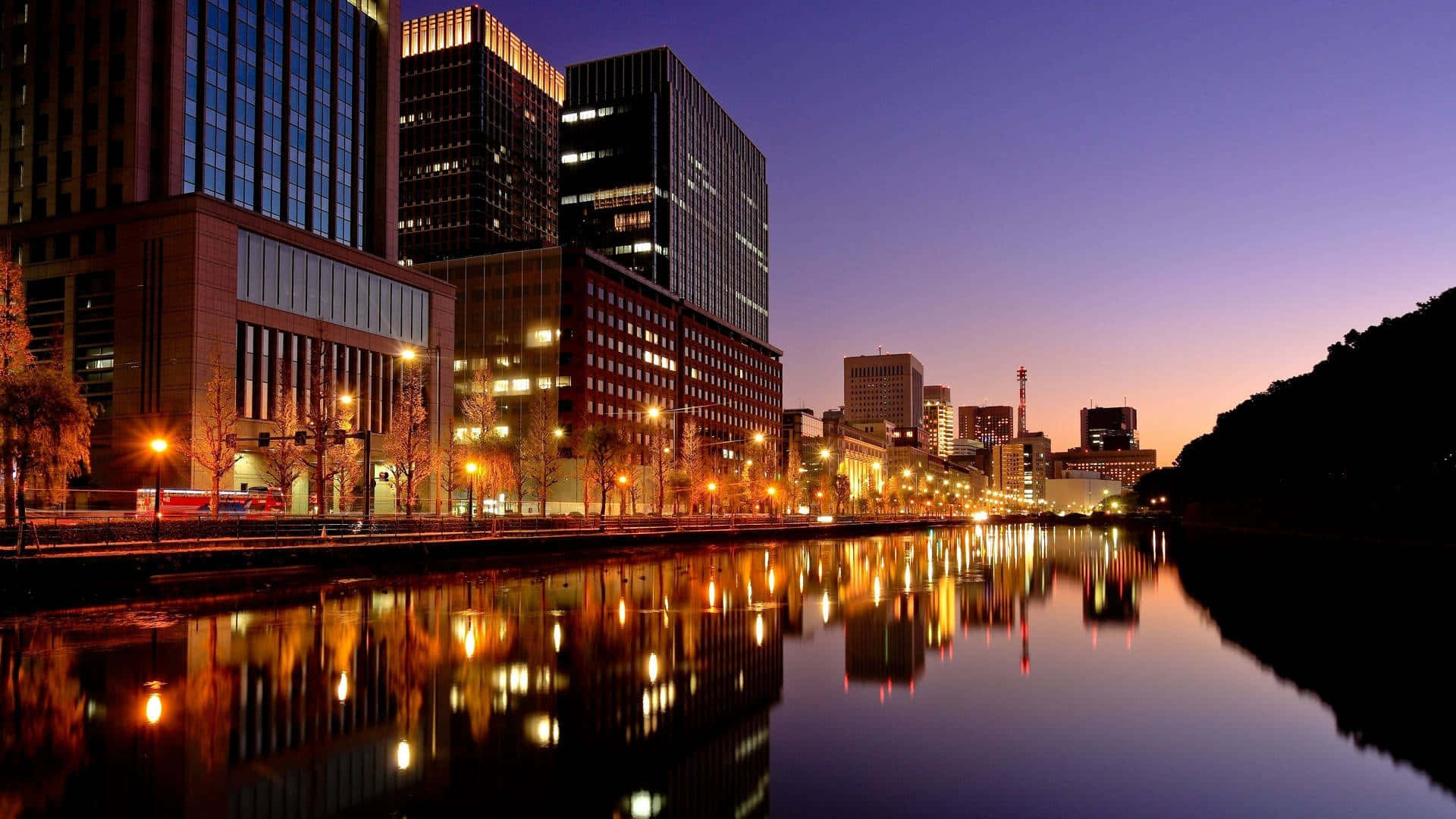 東京 桌布 壁紙 — 黃昏時分的城市天際線，溫暖的路燈和建築物的倒影在平靜的水面上閃爍。