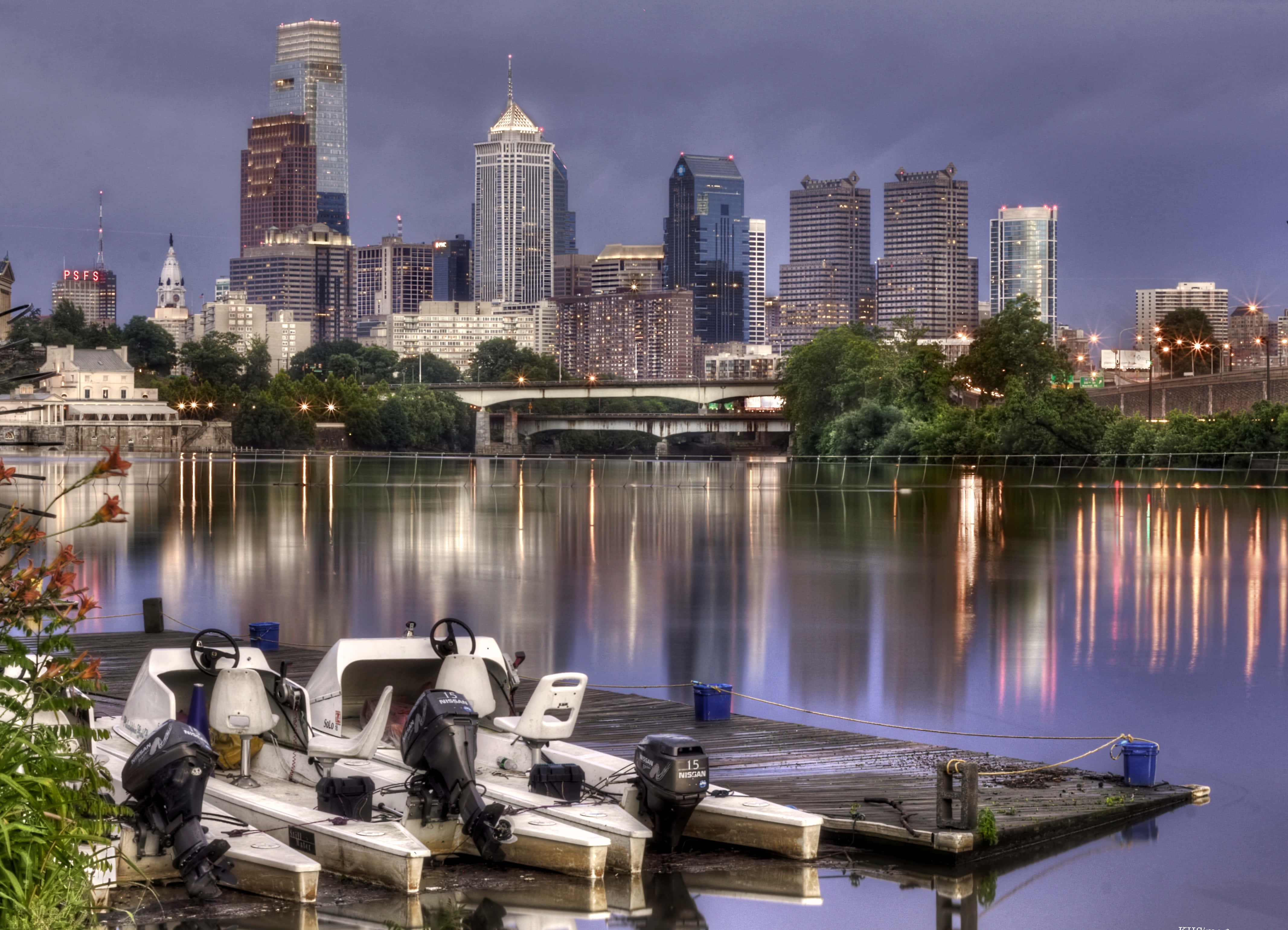 Philly 桌布 壁紙 — 黃昏時分的城市天際線，燈火通明的摩天大樓倒映在平靜的河水上，旁邊停泊著船隻。
