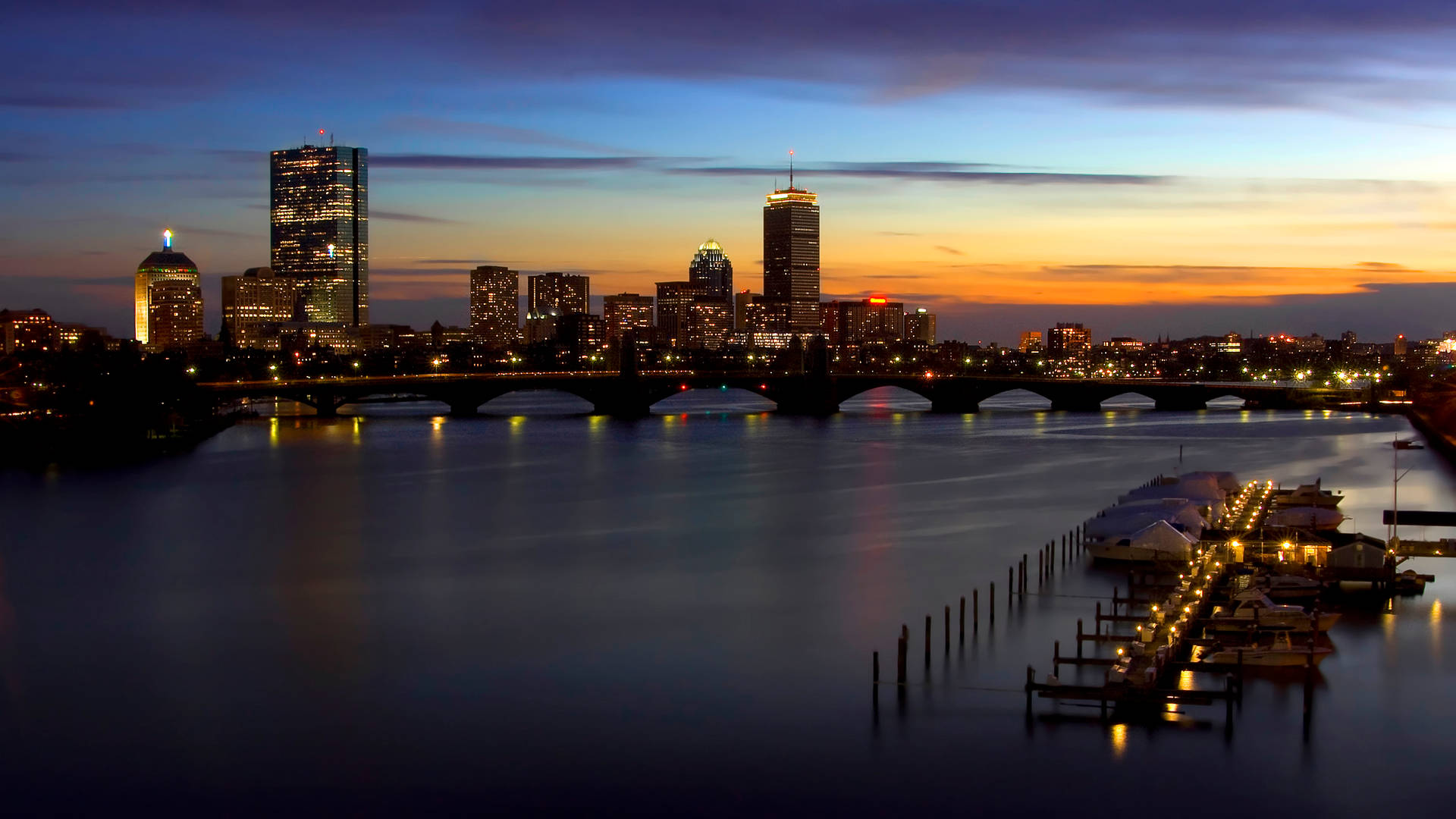New Orleans 桌布 壁紙 — 黃昏時分的城市天際線，燈火通明的摩天大樓和一座橋樑橫跨平靜的水面，反射出溫暖的橙色和冷藍色調。