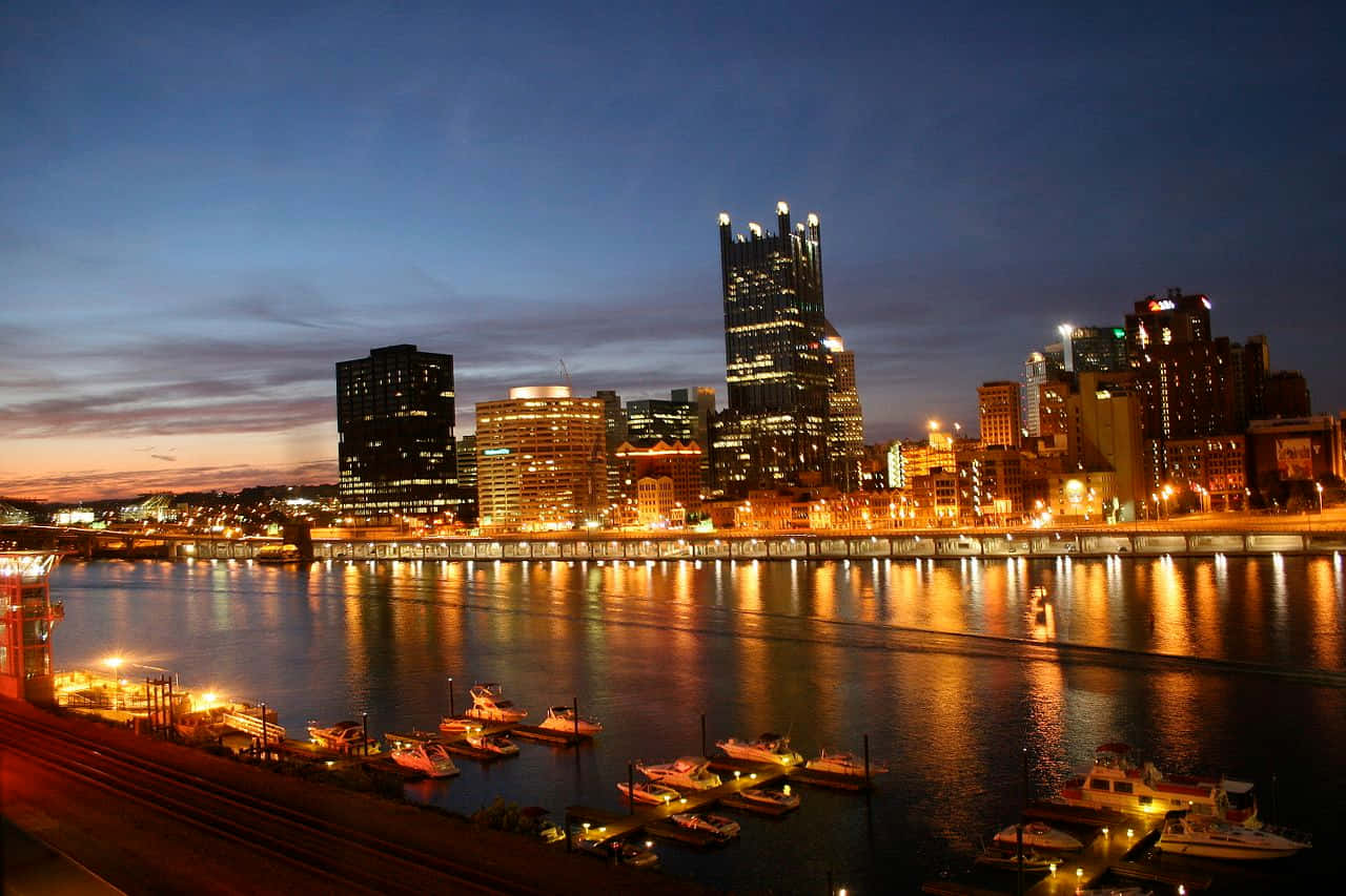 Pittsburgh Skyline 桌布 壁紙 — 黃昏時分的城市天際線，發光的摩天大樓倒映在停泊船隻旁的平靜水面上。