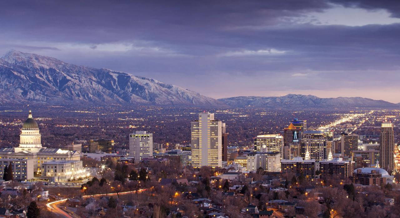Salt Lake City 桌布 壁紙 — 黃昏時分的城市天際線，積雪的山脈在發光的摩天大樓和路燈後面若隱若現。