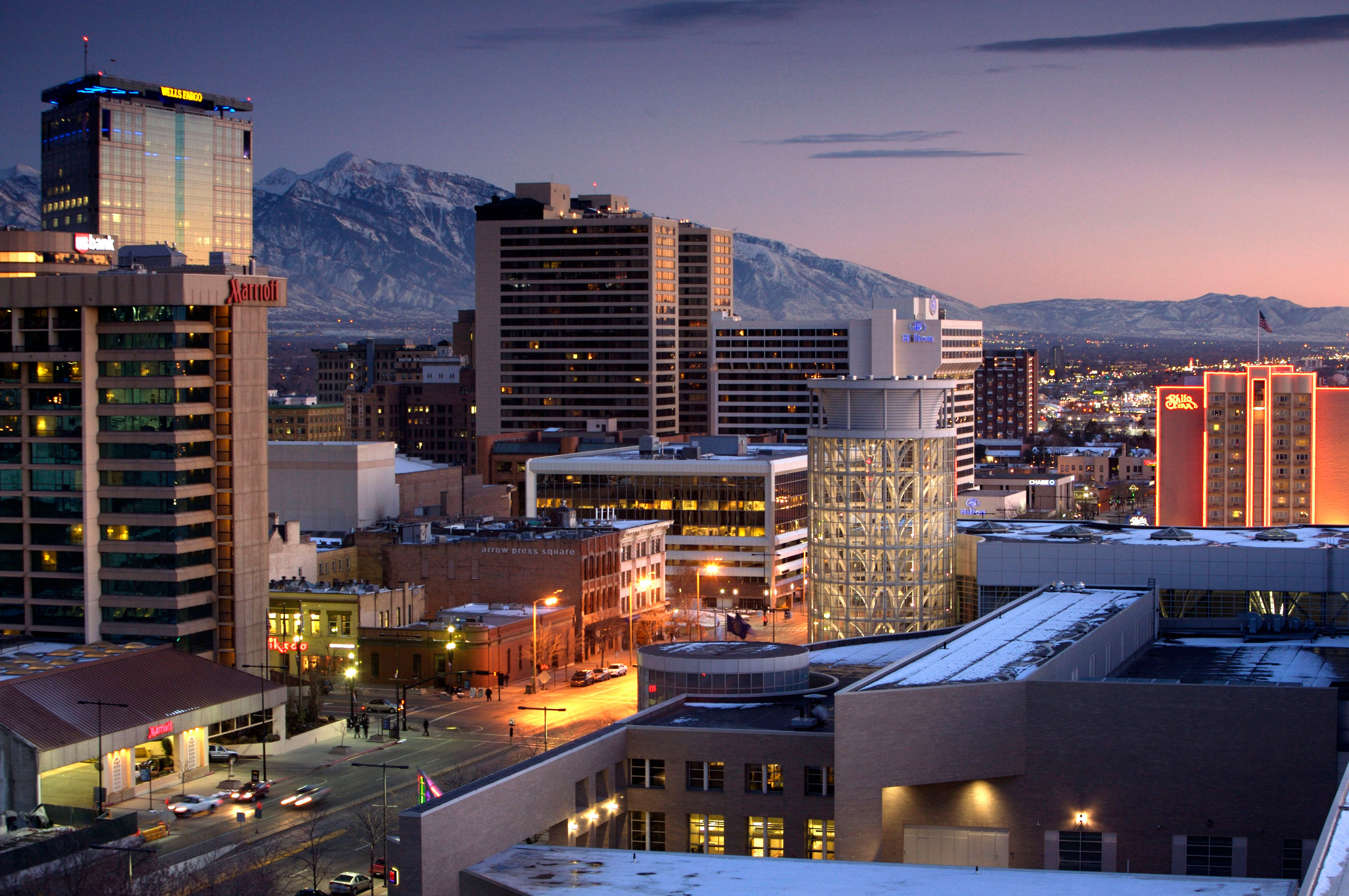 Salt Lake City 桌布 壁紙 — 黃昏時分的城市天際線，積雪的山脈在燈火通明的摩天大樓和街道後方閃耀。