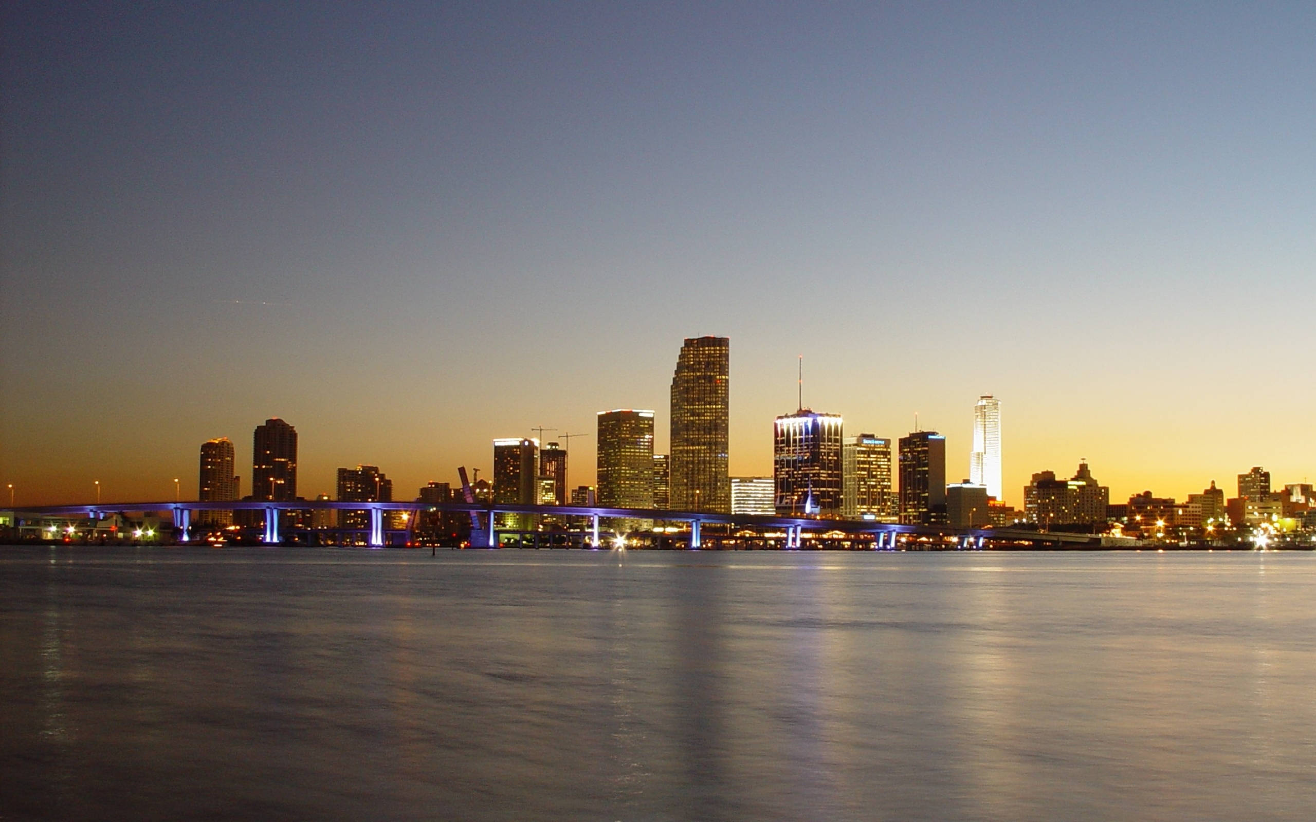 Miami 桌布 壁紙 — 黃昏時分的城市天際線，閃爍的摩天大樓和亮燈的橋樑倒映在平靜的水面上。