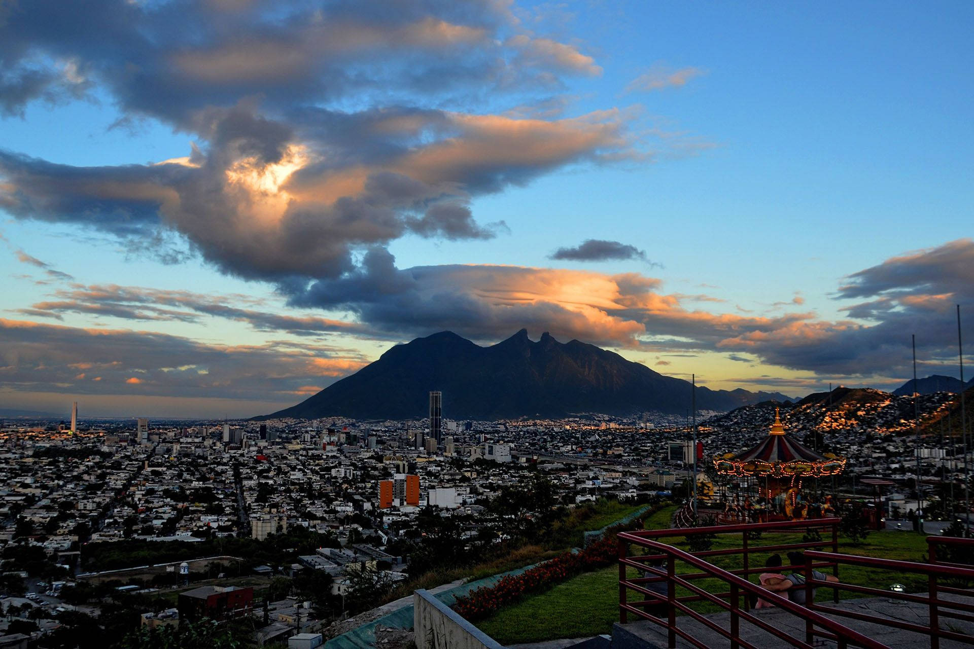 Monterrey 桌布 壁紙 — 黃昏時分的城市景觀，雄偉的山脈輪廓和前景中閃閃發光的旋轉木馬。