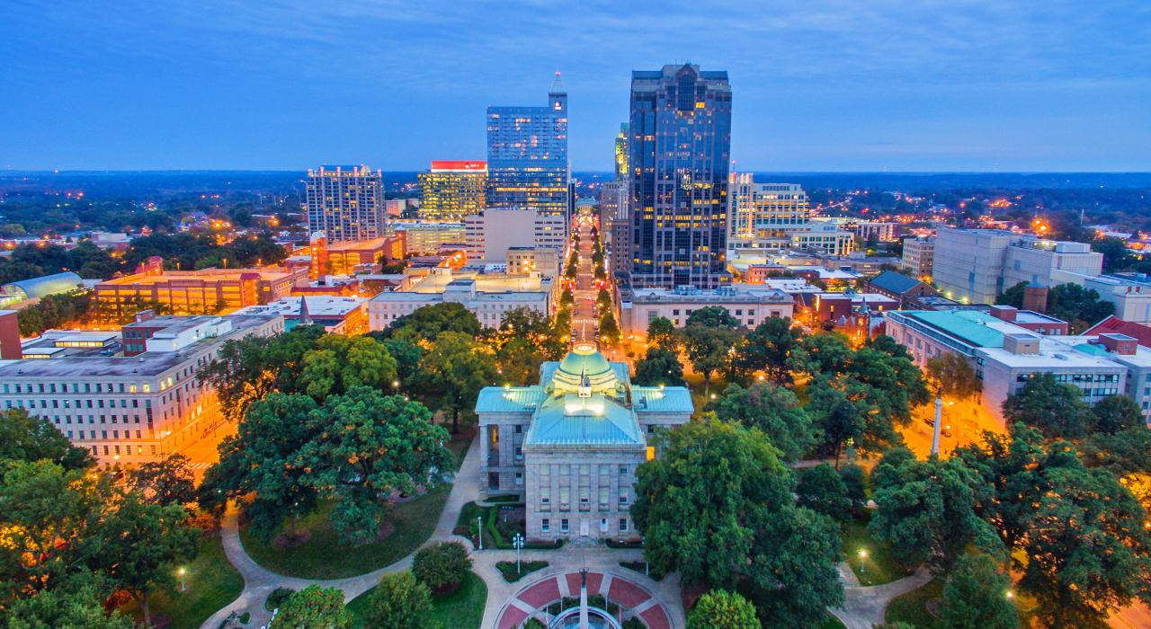 North Carolina 桌布 壁紙 — 黃昏時分的城市景觀鳥瞰圖，燈火通明的摩天大樓和中央圓頂建築，周圍環繞著茂密的綠樹。