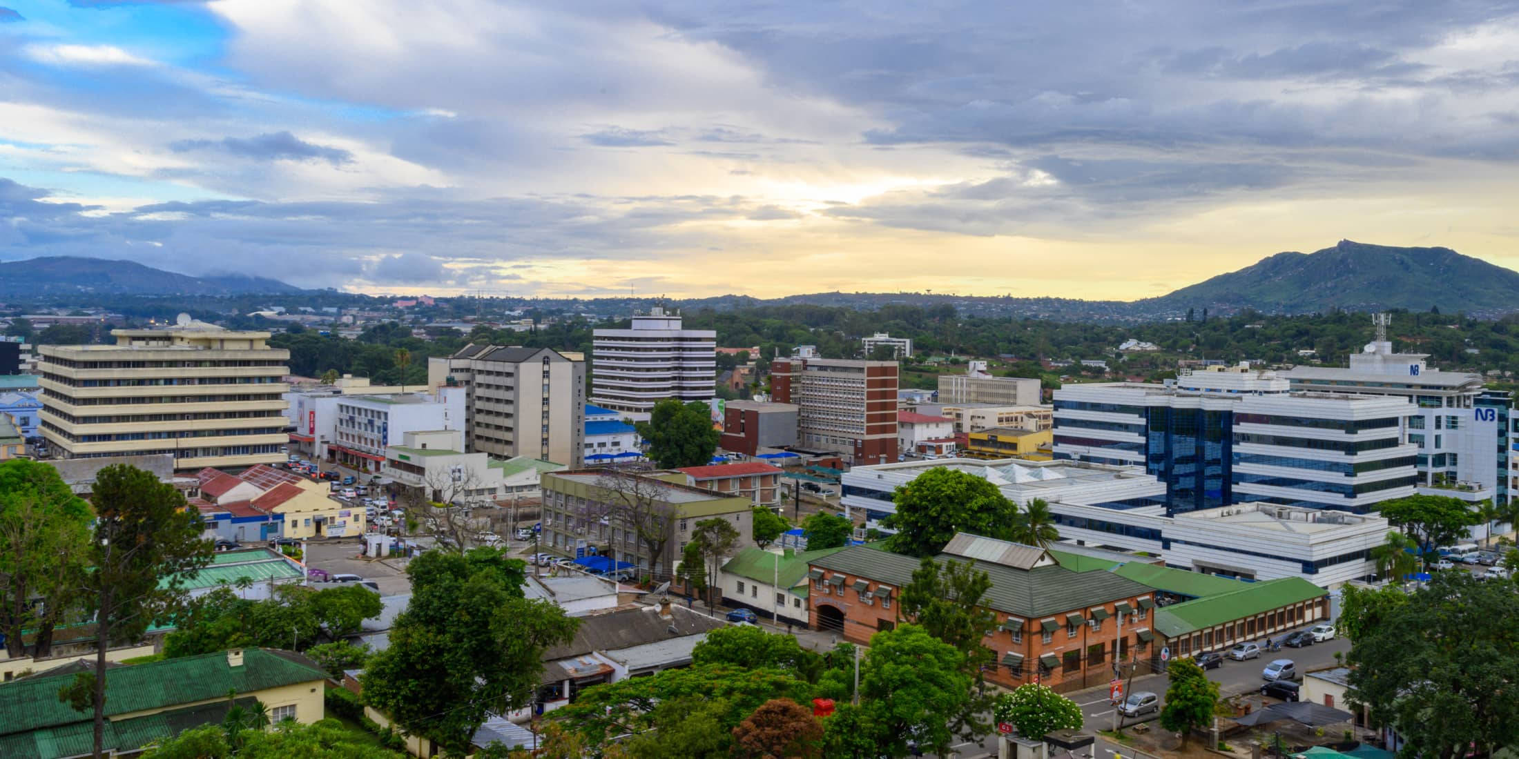 Malawi 桌布 壁紙 — 黃昏時分的城市景觀鳥瞰圖，現代與傳統建築錯落有致，坐落在茂密的綠樹之中，天空多雲。