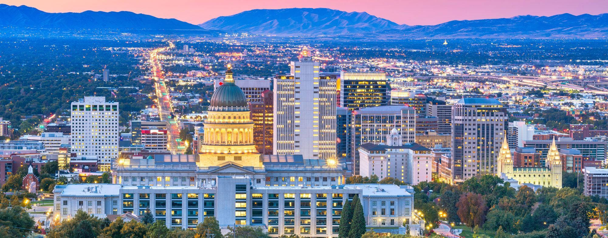 Salt Lake City 桌布 壁紙 — 黃昏時分的城市景觀鳥瞰圖，粉紅色和紫色的天空下，建築物燈火通明，遠山隱約可見。