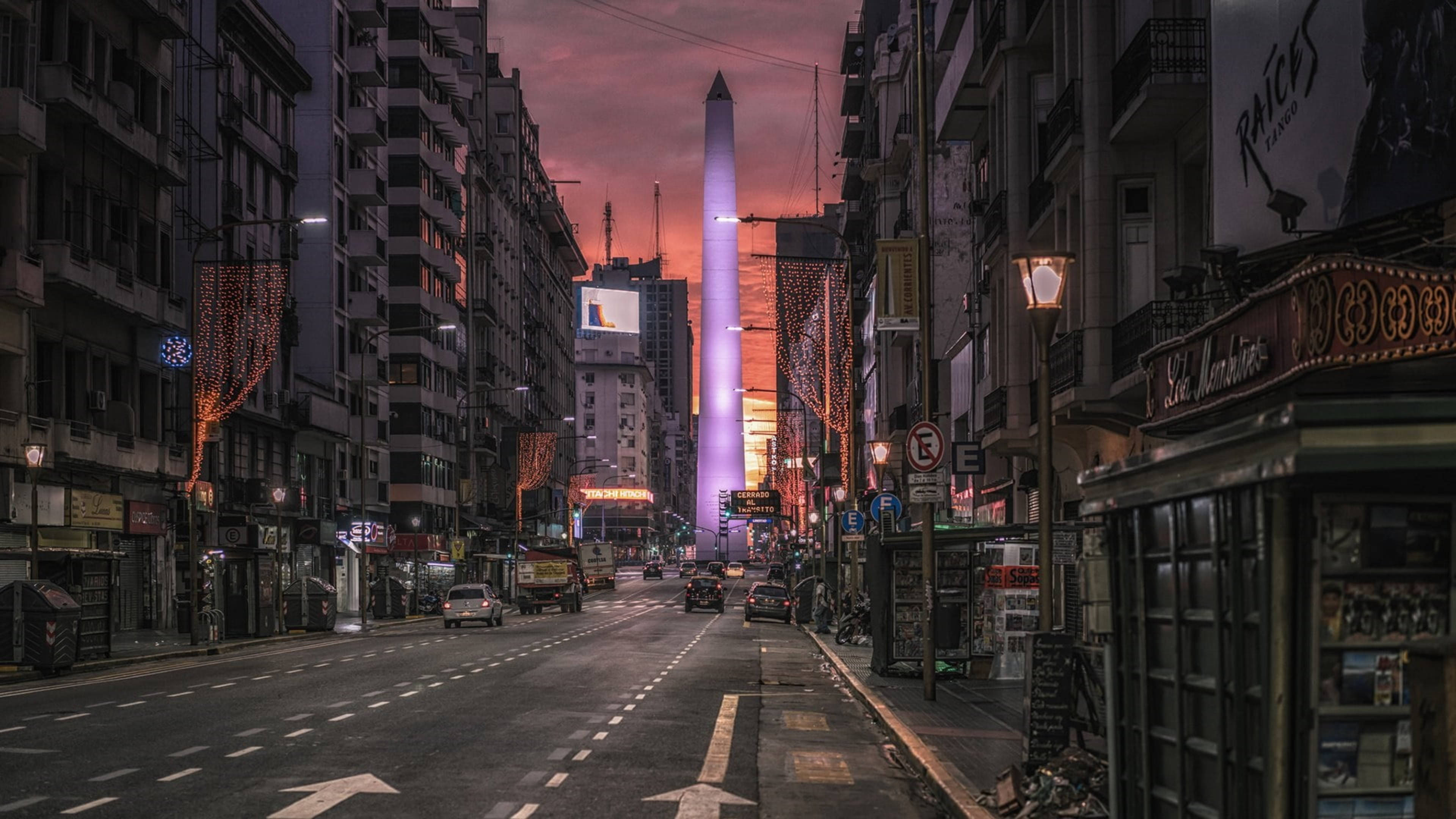 Argentina 桌布 壁紙 — 黃昏時分的城市街道，一座發光的方尖碑高聳於建築物之上，建築物之間掛滿節日燈飾。