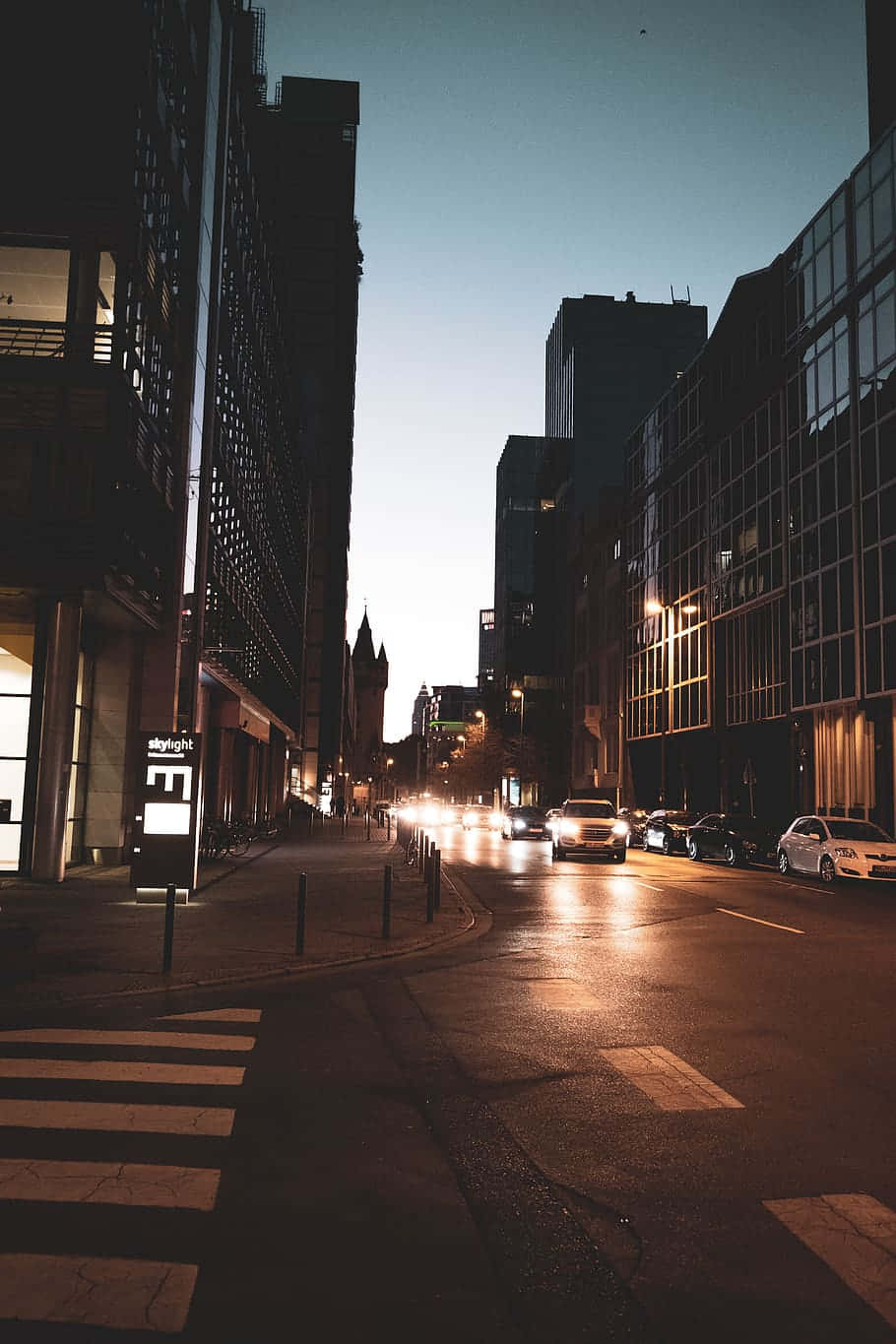 Night Street 桌布 壁紙 — 黃昏時分的城市街道，發光的車頭燈在濕漉漉的路面上反射，映照在高聳的玻璃和混凝土建築之間。