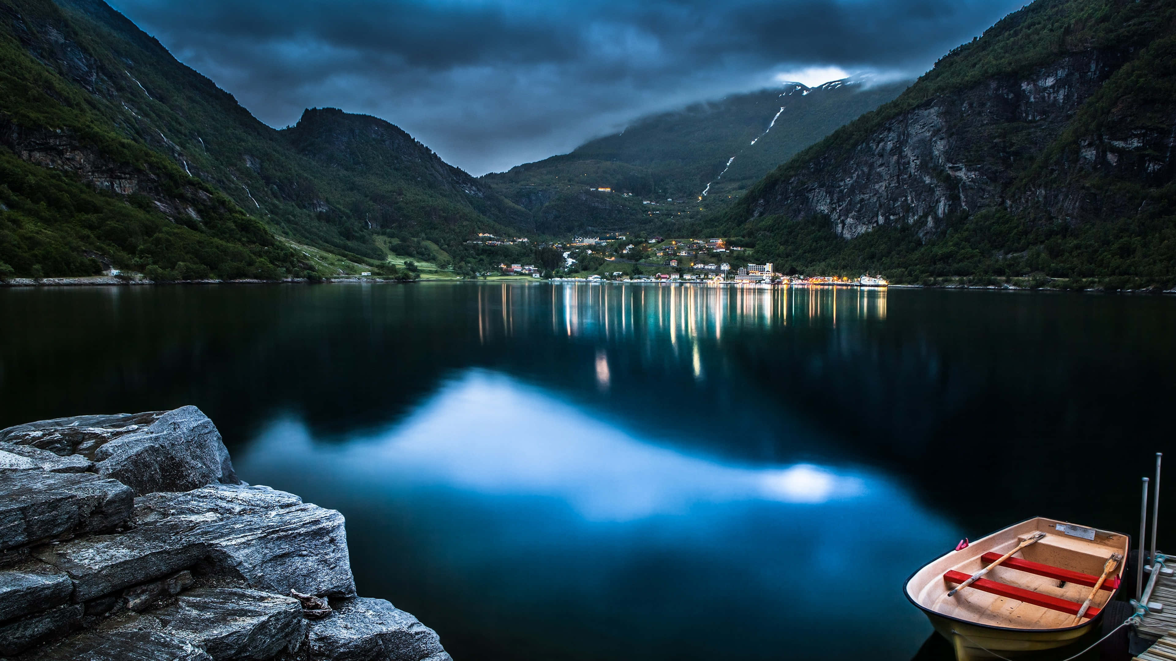 Beautiful Mountain Lake 桌布 壁紙 — 黃昏時分的寧靜峽灣，一艘木船停泊在岩石附近，村莊的燈光在平靜的水面上閃爍倒映。