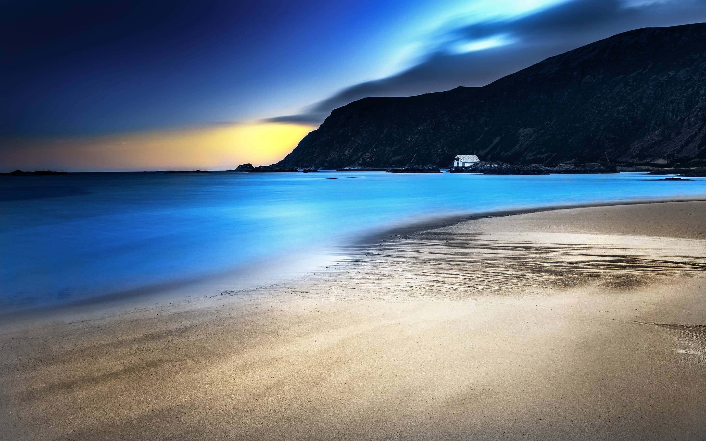 Beach At Night 桌布 壁紙 — 黃昏時分的寧靜海灘，平滑的藍色水流過金色沙灘，一間孤獨的房子依偎在黑暗的懸崖邊。